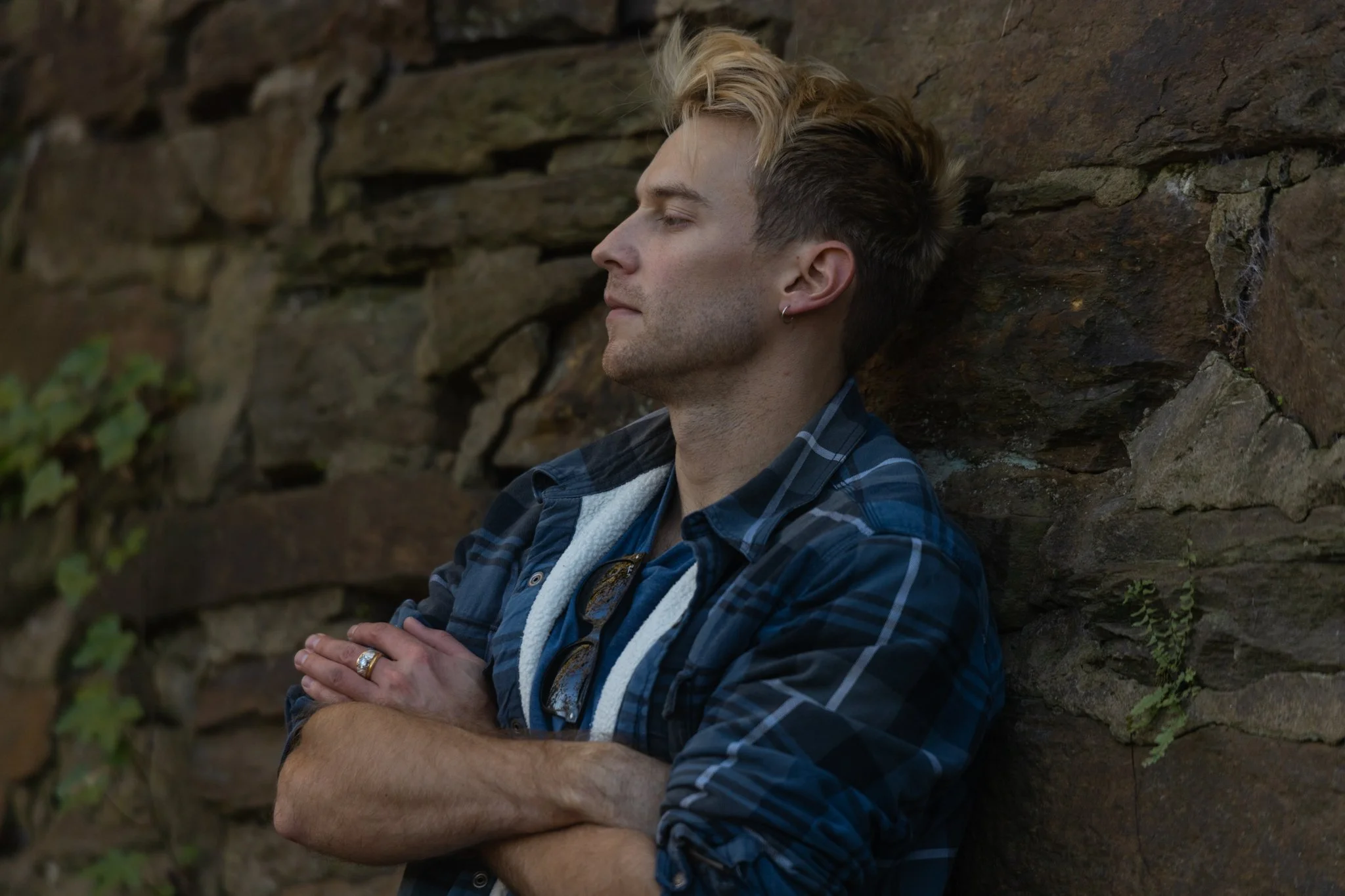 A young man with blond and dark hair, wearing a blue plaid shirt and sunglasses hanging from his collar, stands with his arms crossed and eyes closed against a stone wall.