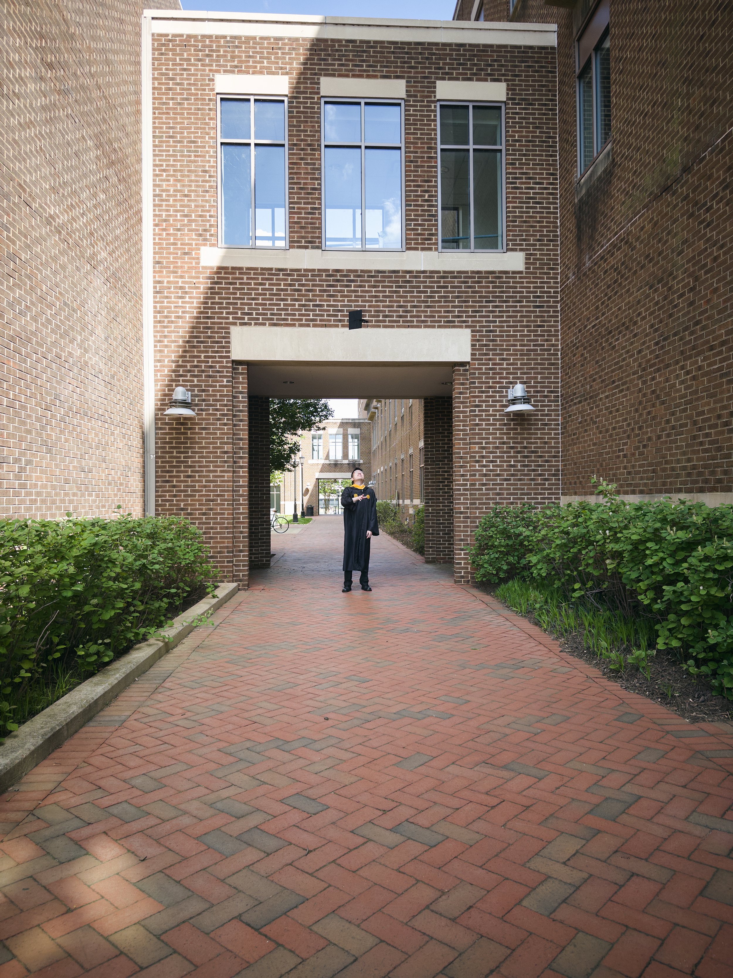 A person in a graduation gown and cap standing under a brick archway on an outdoor pathway.