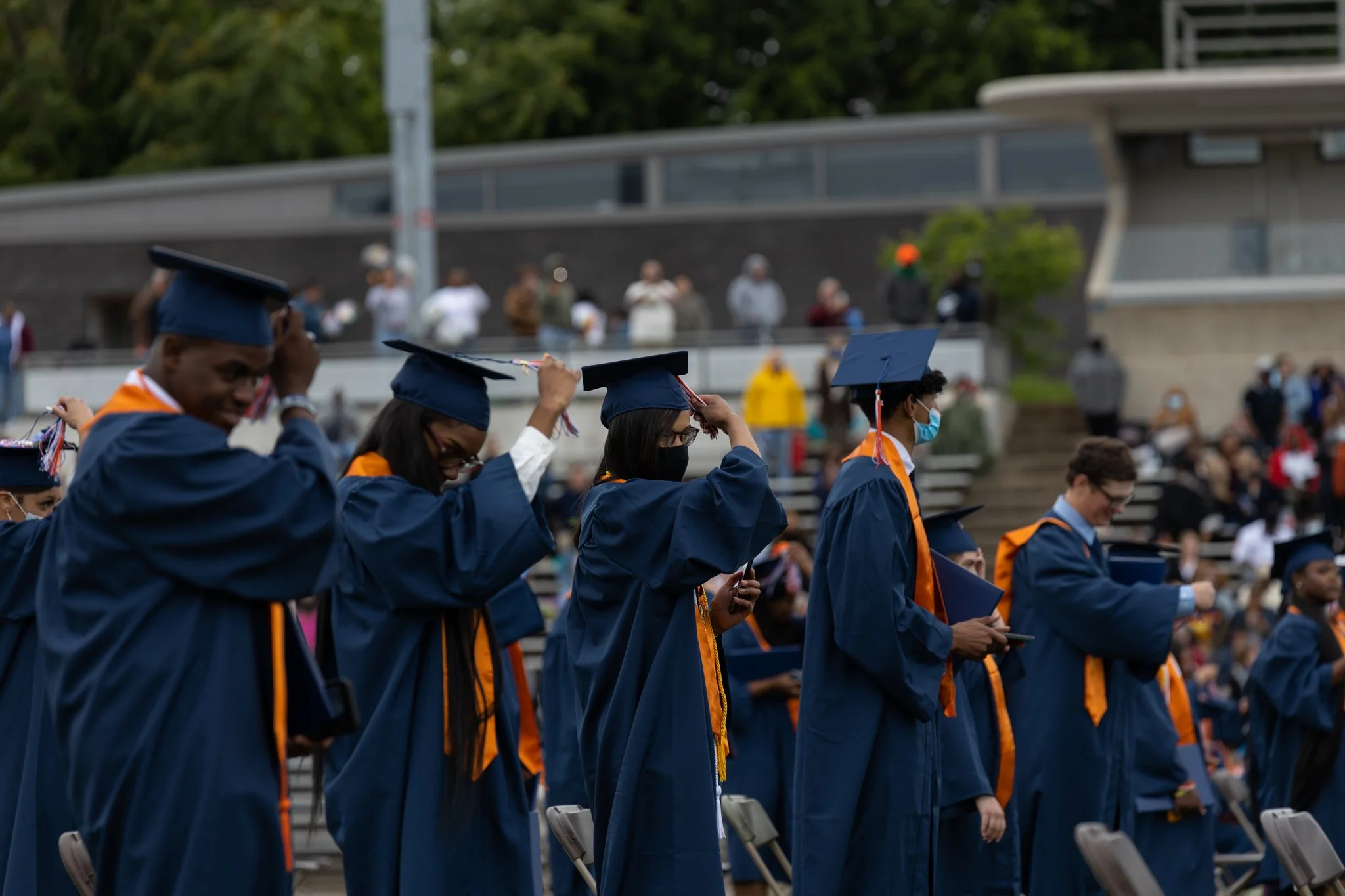 Group of graduates in blue caps and gowns at an outdoor graduation ceremony, adjusting their caps, some wearing face masks.