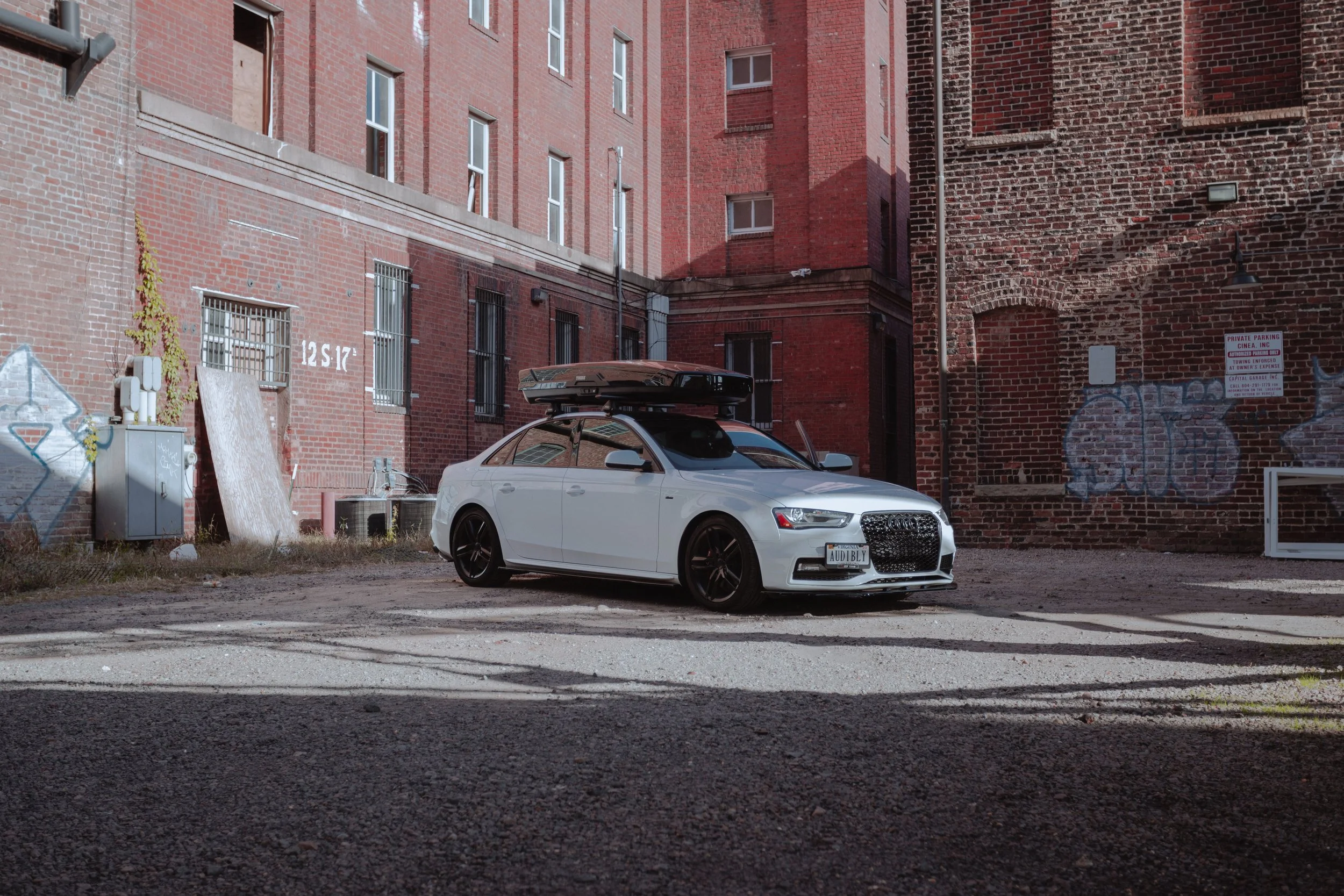 A white Audi sedan with a black roof box parked in an alley between red brick buildings, with graffiti on the wall and a sign indicating private parking.