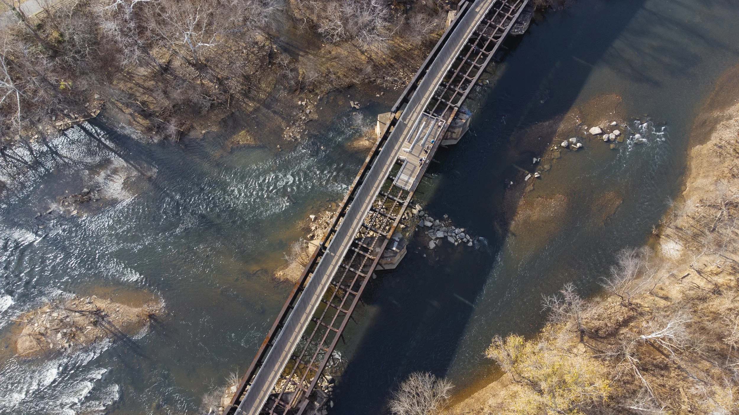 Aerial view of a bridge crossing over a river with rocky banks and leafless trees on the sides.
