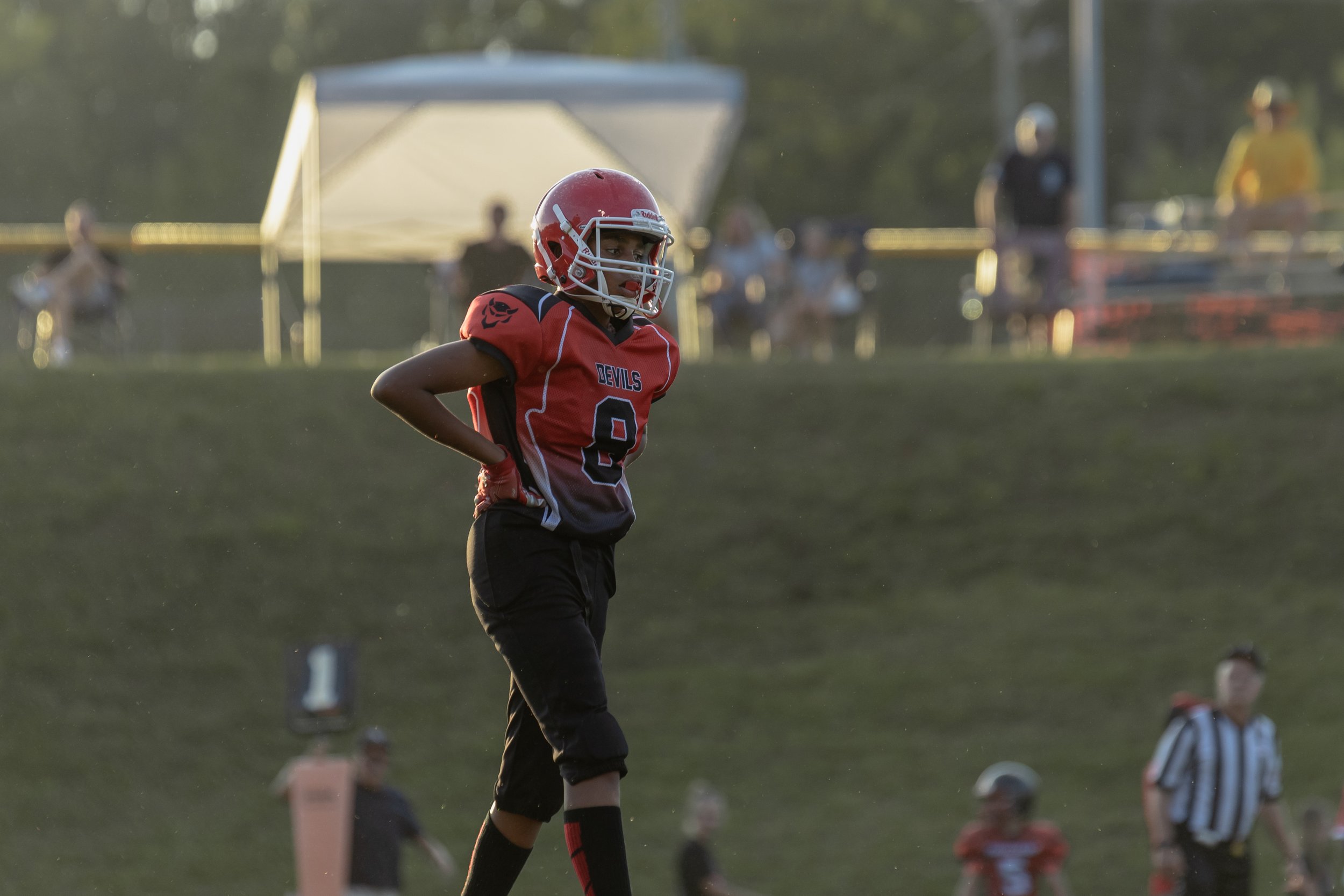 A young football player standing on the field in a red and black uniform with a helmet, during a game. The background shows spectators and officials.