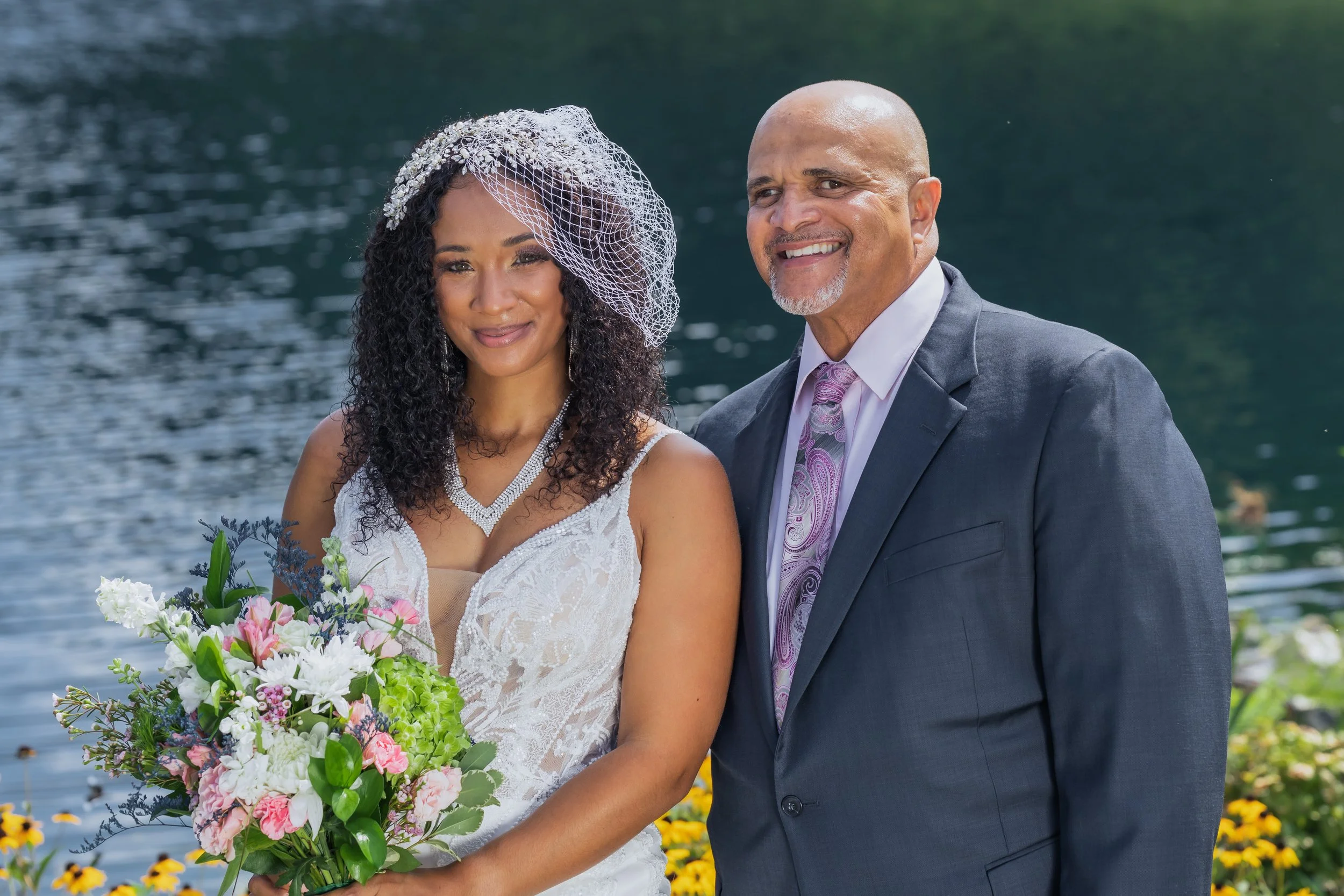 A bride and an older man, likely her father, posing outdoors near a lake with trees and flowers in the background. The bride is wearing a white wedding dress, veil, and holding a bouquet of flowers. The man is wearing a dark suit and a patterned tie.