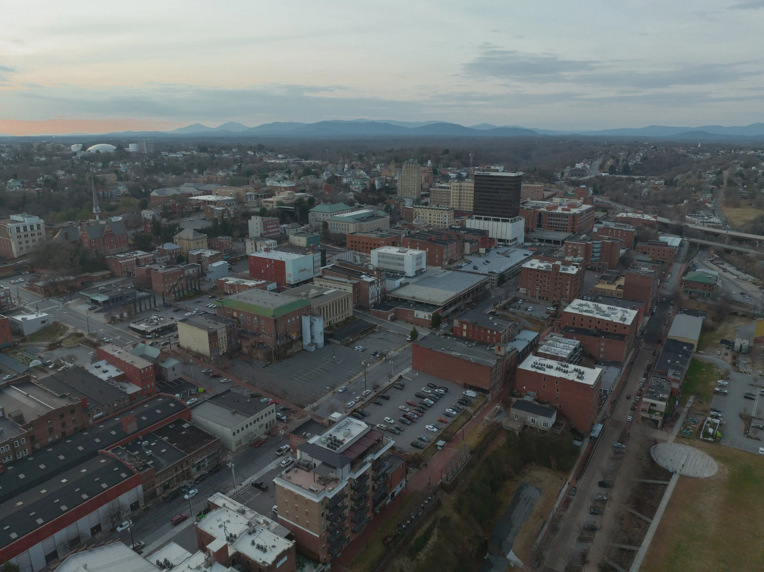Aerial view of a city with a mix of historic and modern buildings, parking lots, and streets, with mountains in the background during sunset.