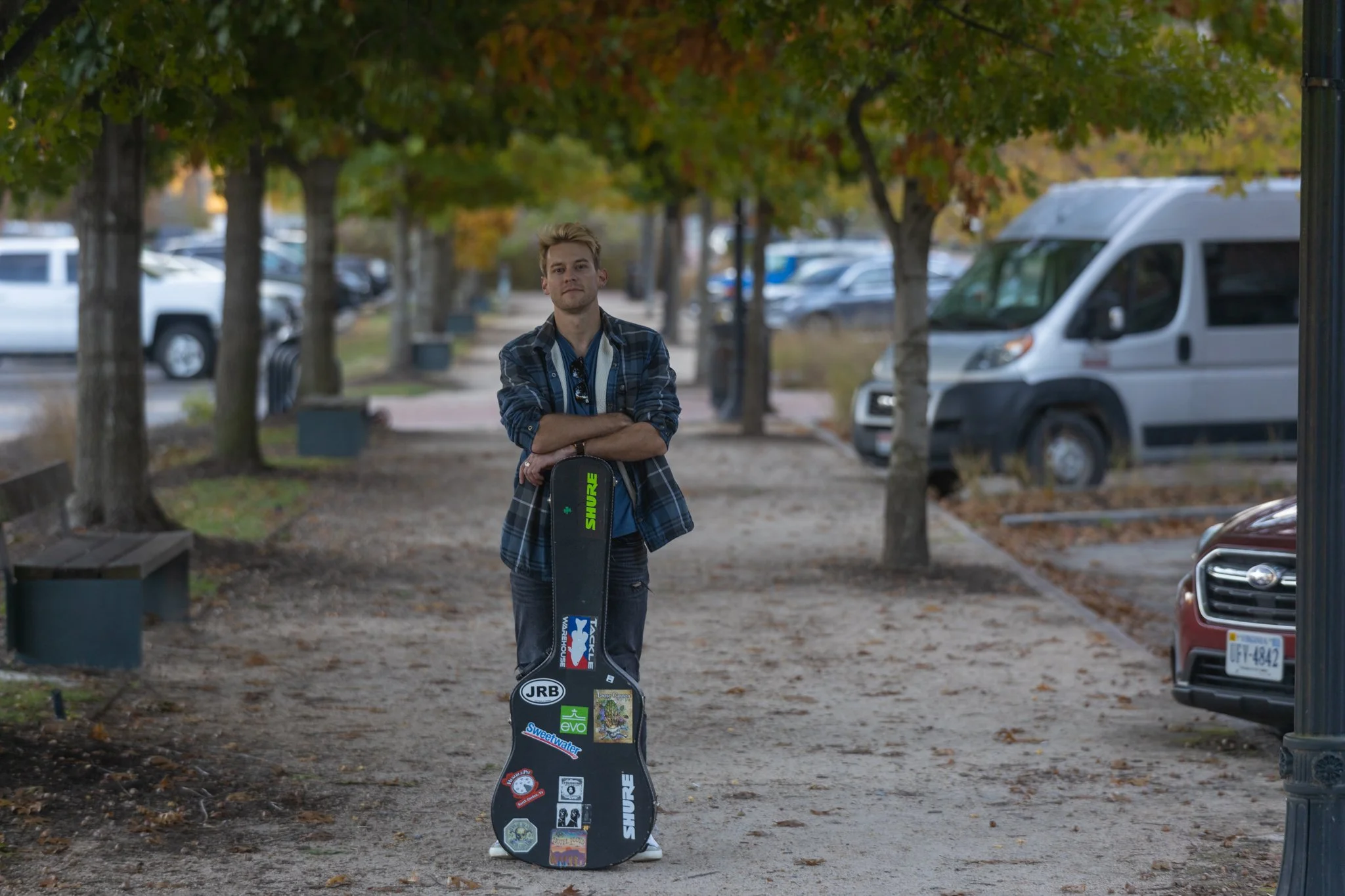 A young man standing on a tree-lined sidewalk with his arms crossed, holding a guitar case covered with stickers. Parked cars are in the background, and the scene suggests a fall setting.