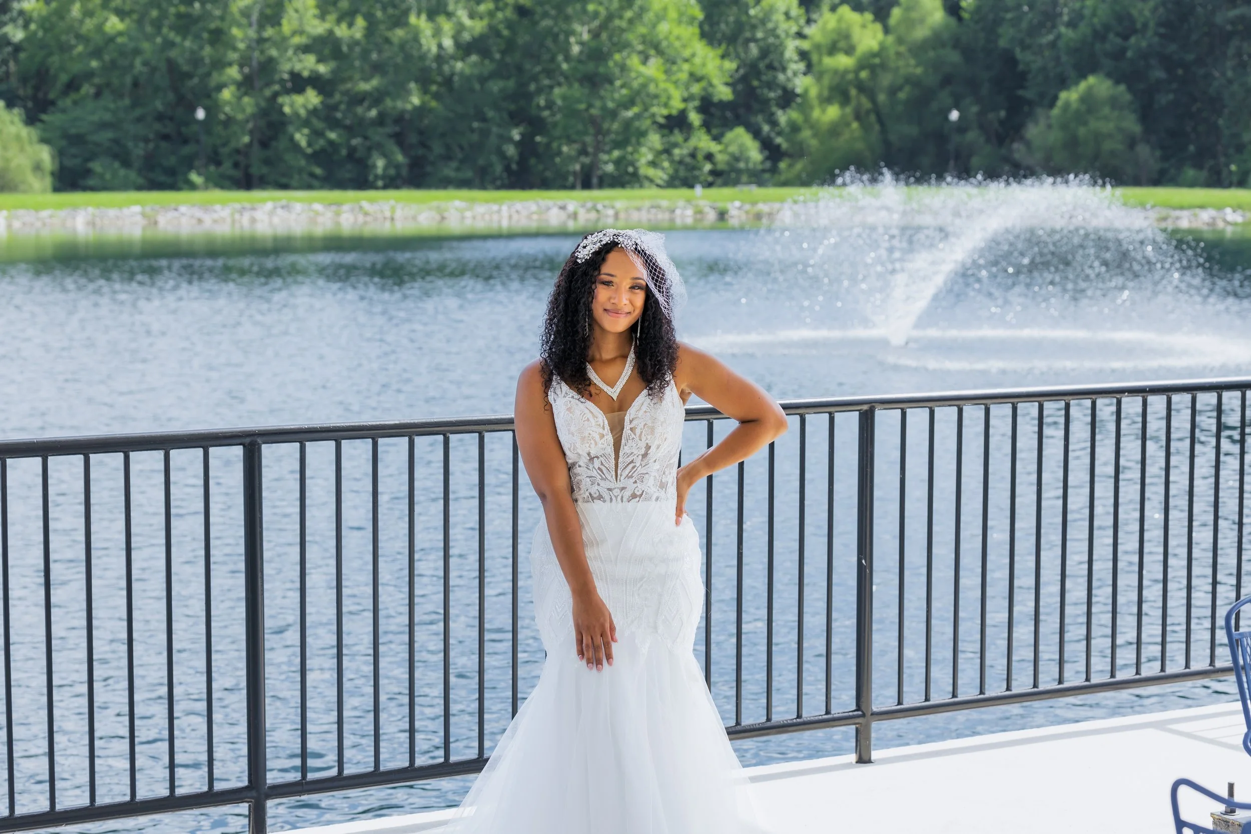 Woman in a white lace wedding dress with veil smiling by a lake with a fountain and green trees in the background.