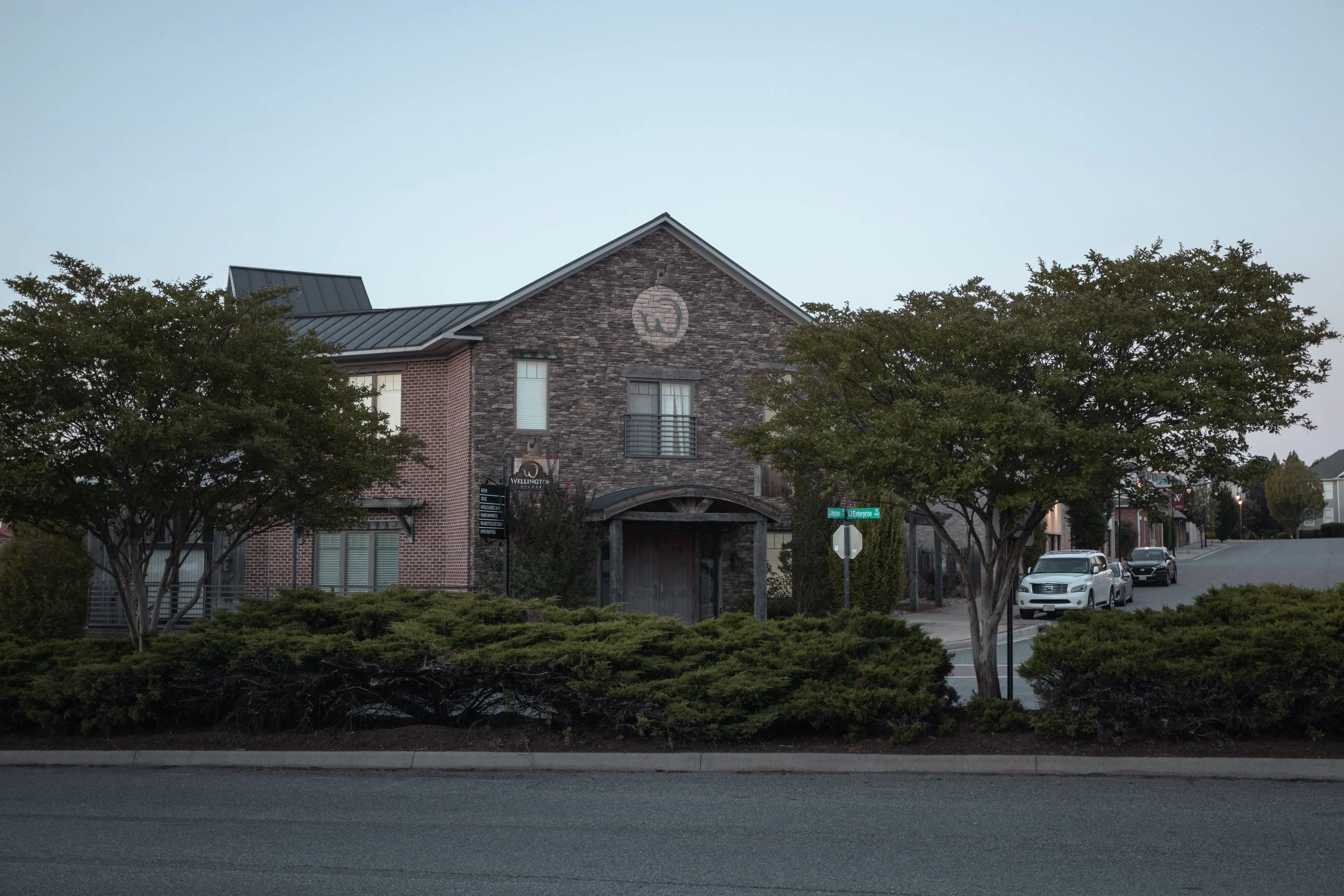 A commercial building with brick and stone facade, trees in the foreground, parked cars to the right, and a street sign that reads 'Wellington' in an urban area.