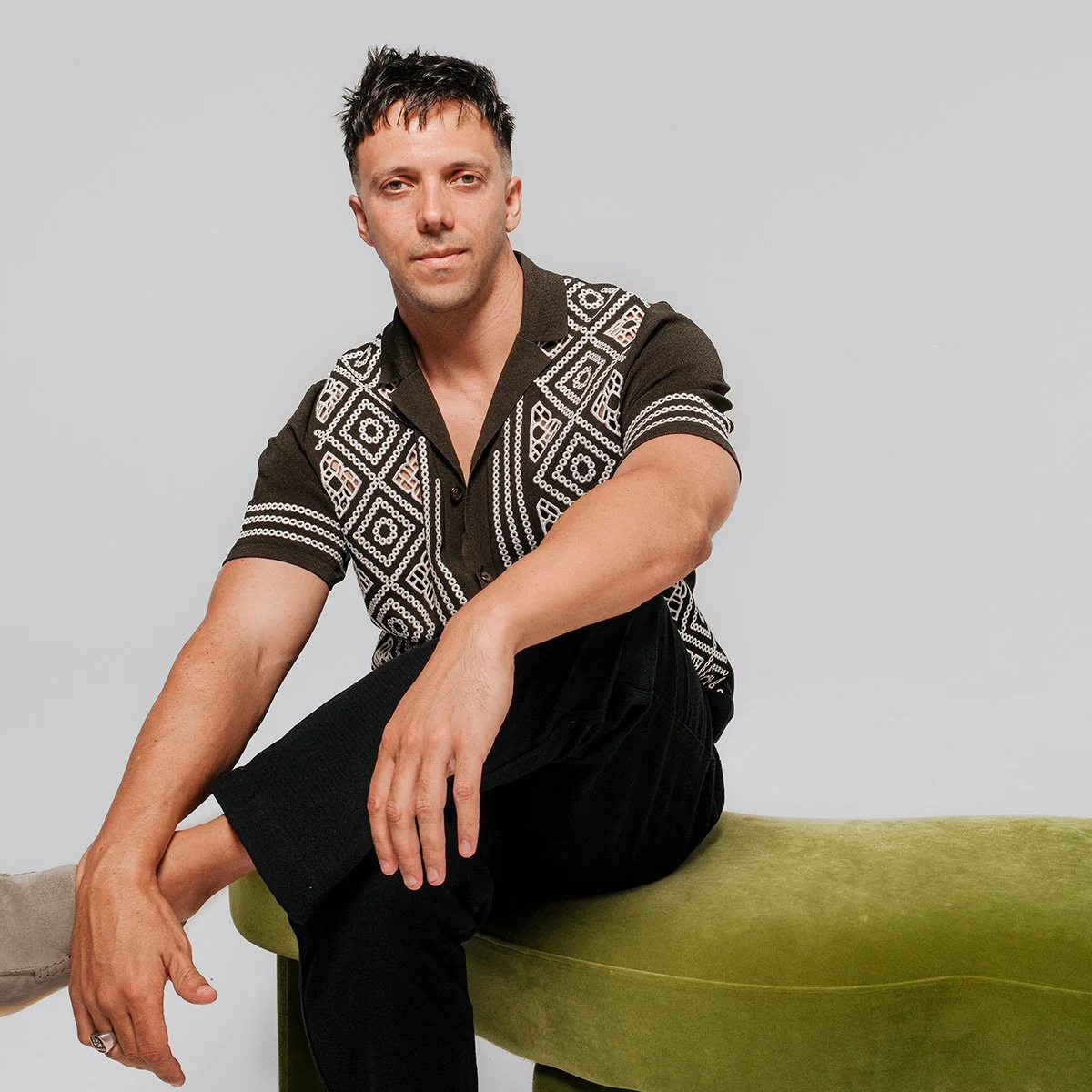 Man sitting on green bench wearing patterned shirt and black pants, against a gray background.