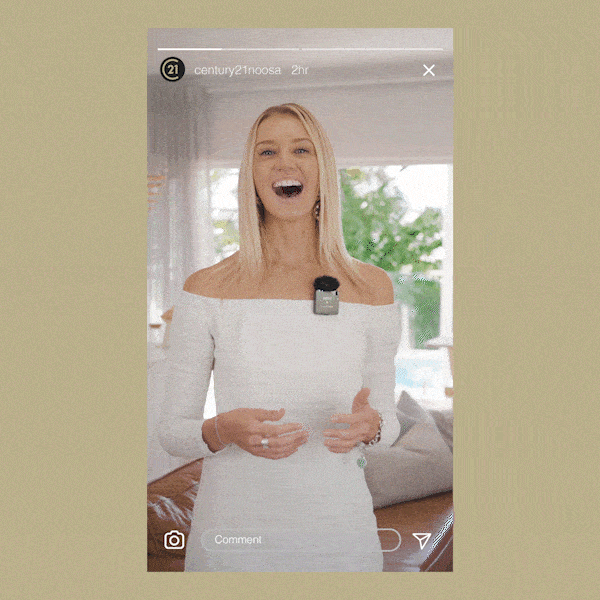 A woman in a white dress smiling, standing indoors with a clip microphone on her dress. The background shows a bright room with a couch and window view.