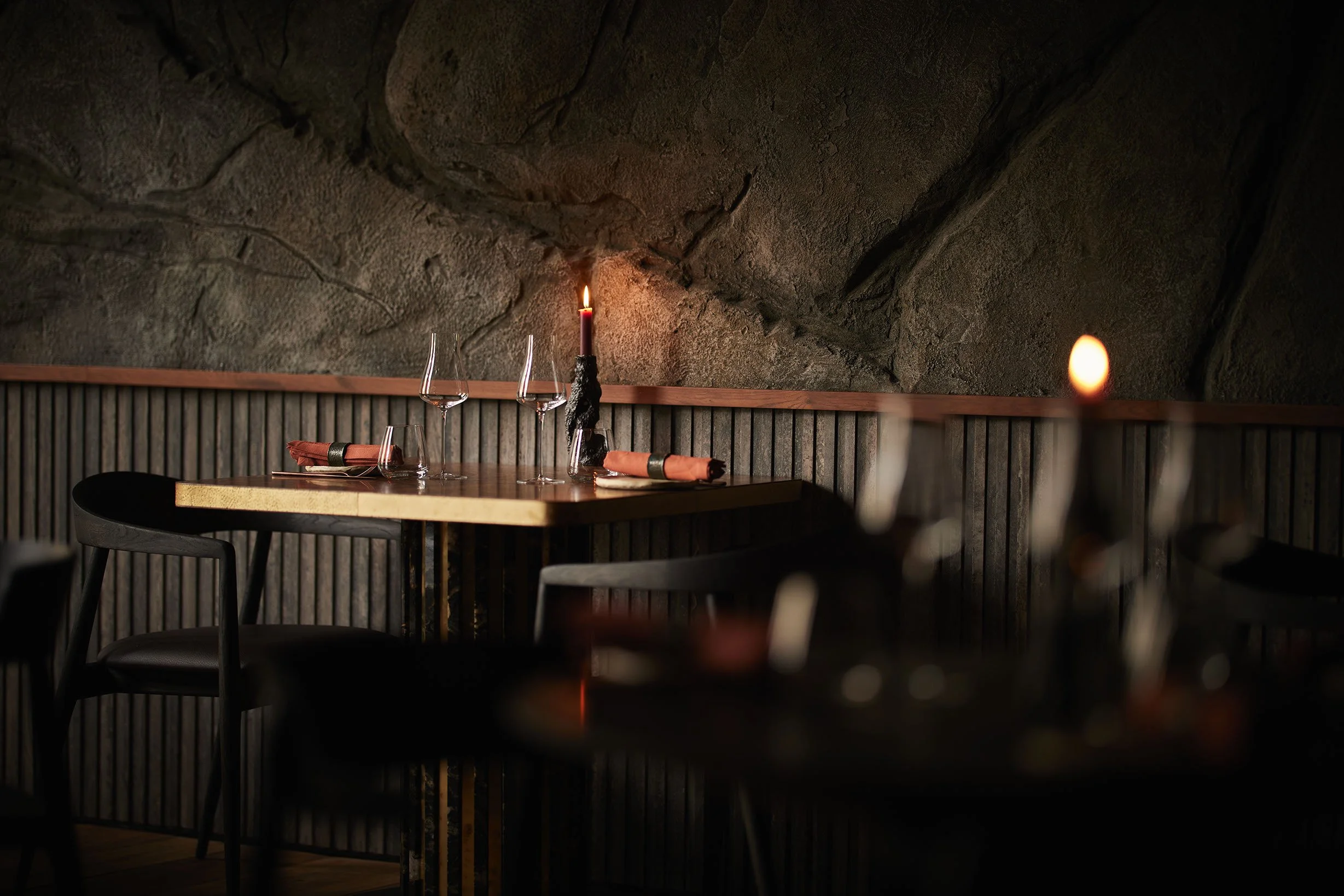 A cozy restaurant setting with a wooden table, wine glasses, napkins, and lit candles against a rock wall background.
