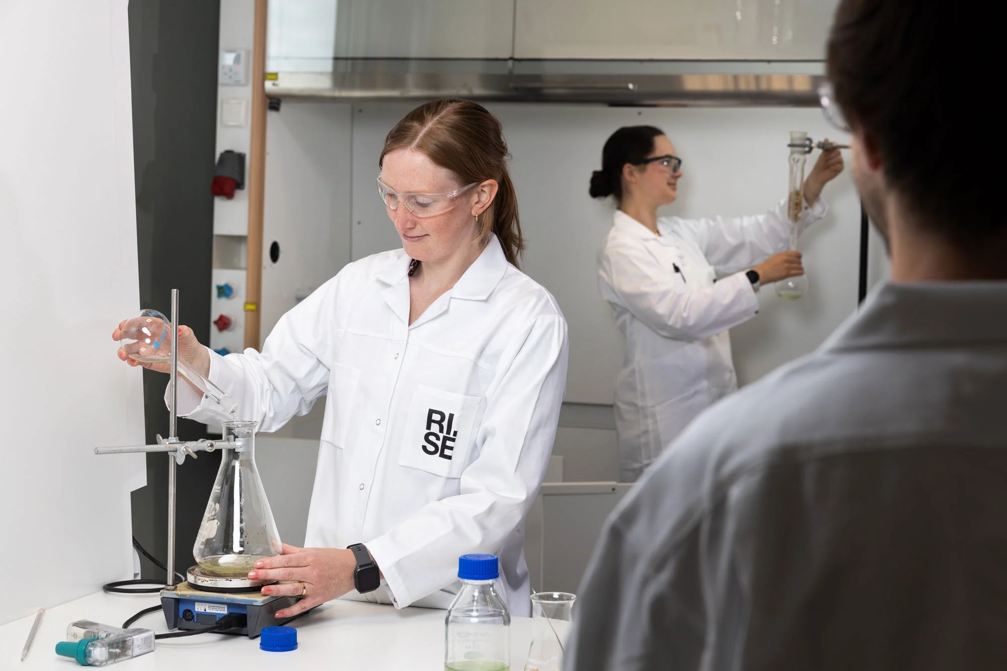Forskare på RISE i labb. En kvinna med skyddsglasögon häller vätska i en behållare.