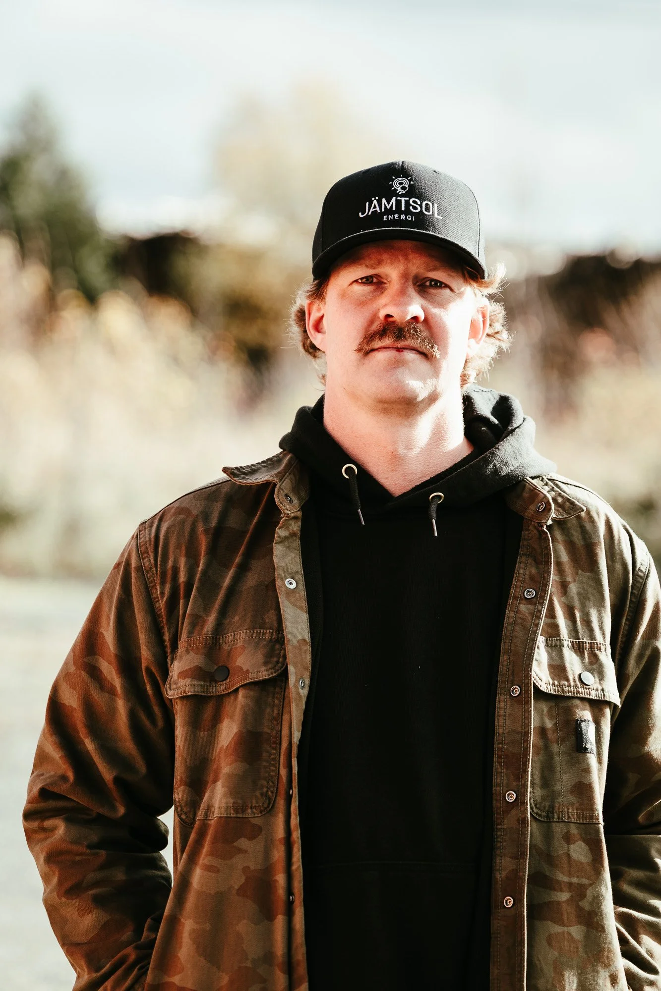 Porträtt på Erik Vareberg på JS El & Metall. Han bär en svart huvtröja och keps, med en kamoflagefärgad jacka över.