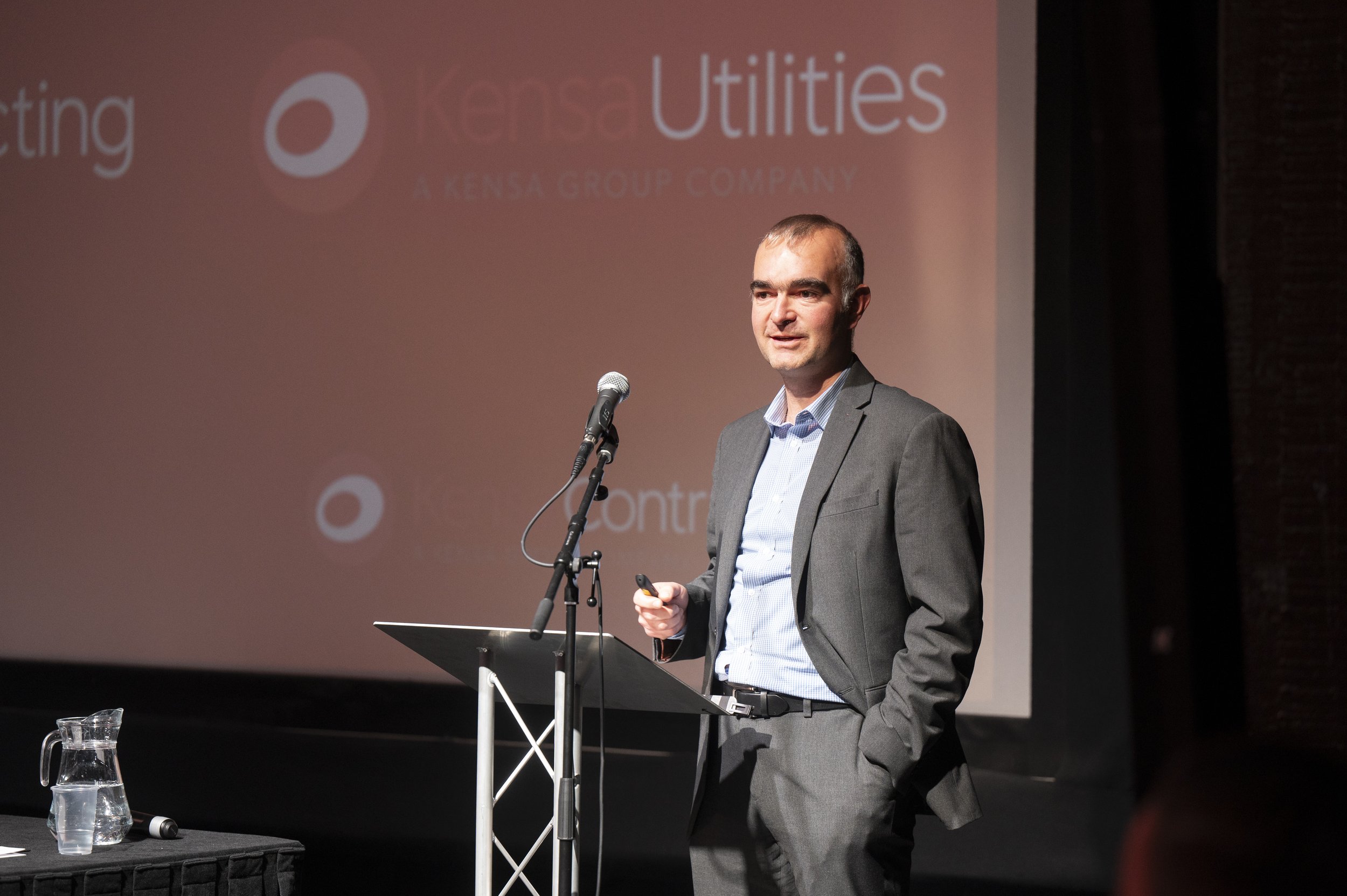 Person in suit speaking at a podium with "Kensa Utilities" on screen in background.