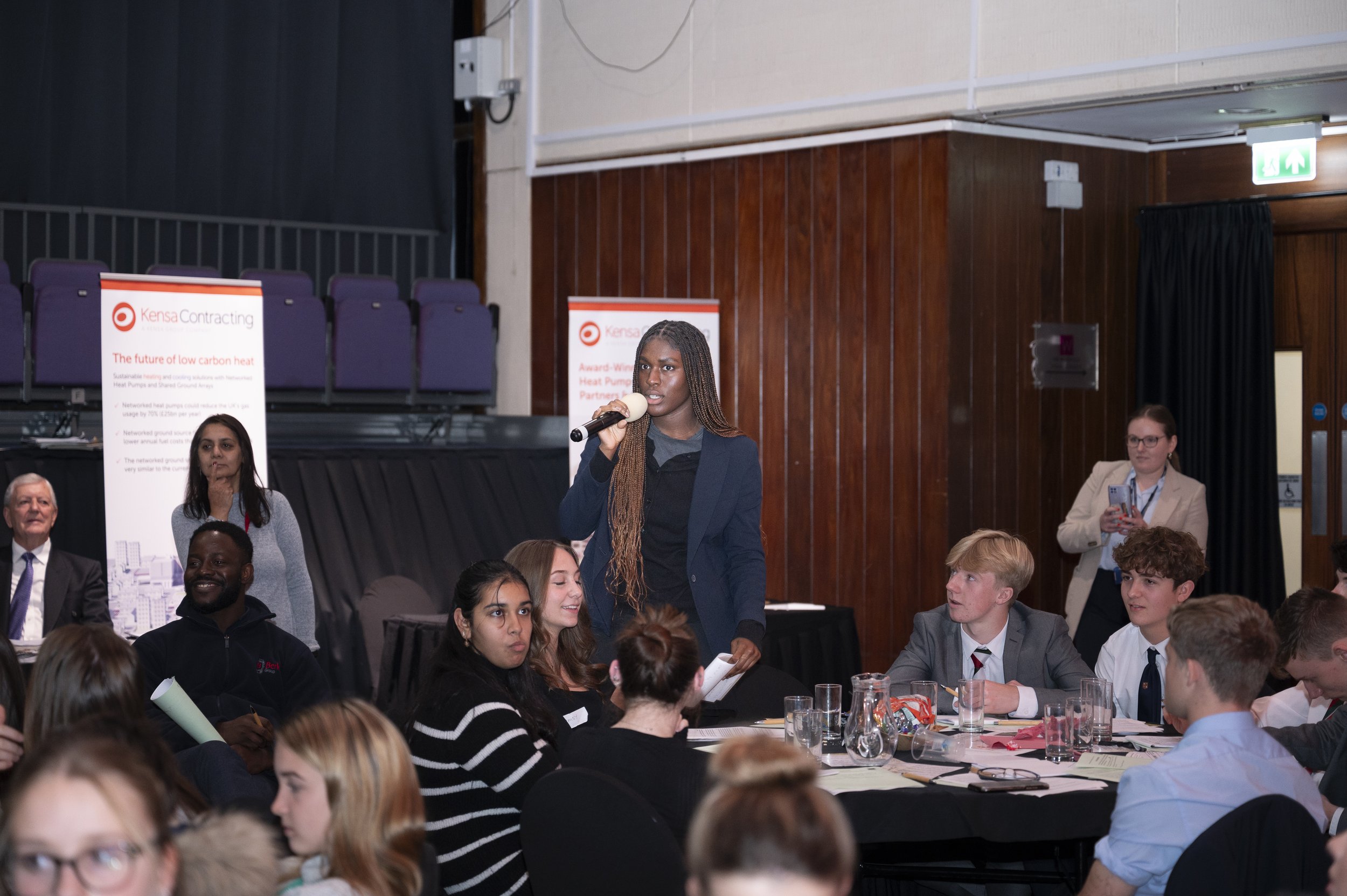 A person speaking into a microphone at a conference or seminar, surrounded by seated attendees. Banners for Kensa Contracting are visible in the background.