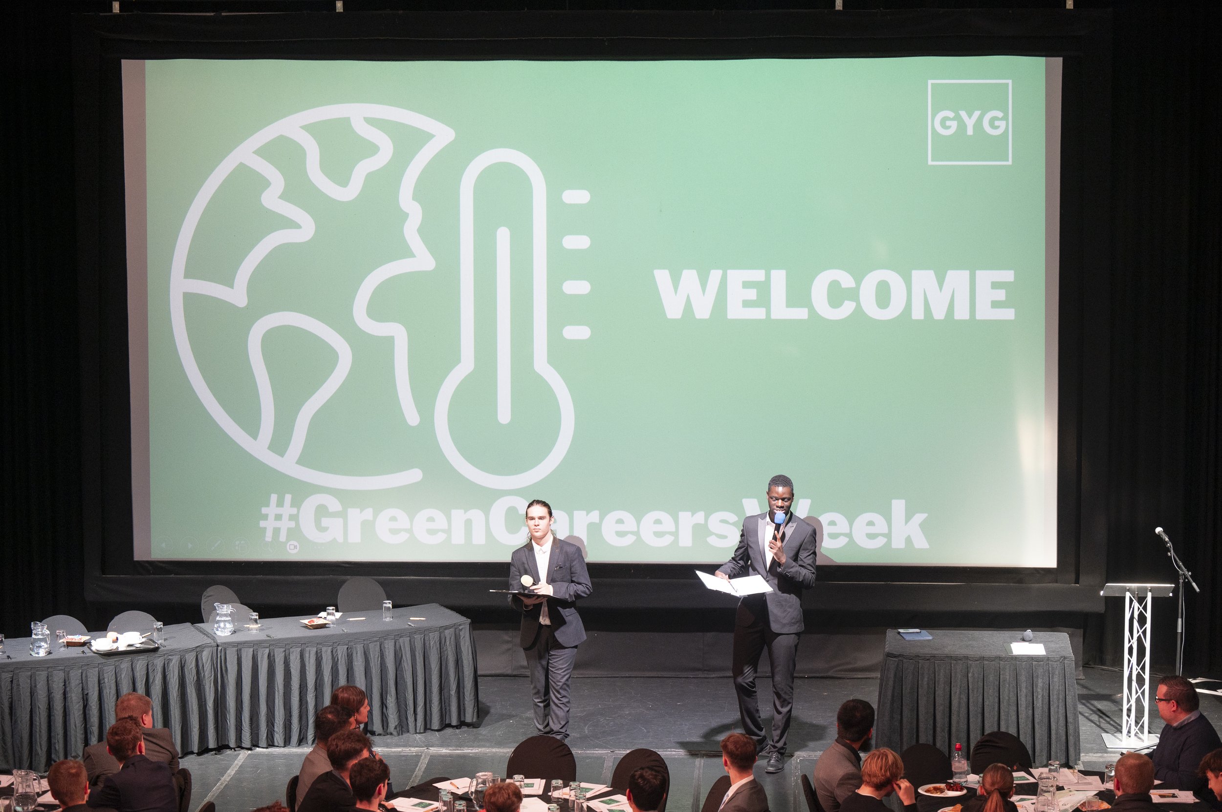 Conference stage with two presenters, large screen displaying "WELCOME" and "#GreenCareersWeek" logo, audience seated at tables.