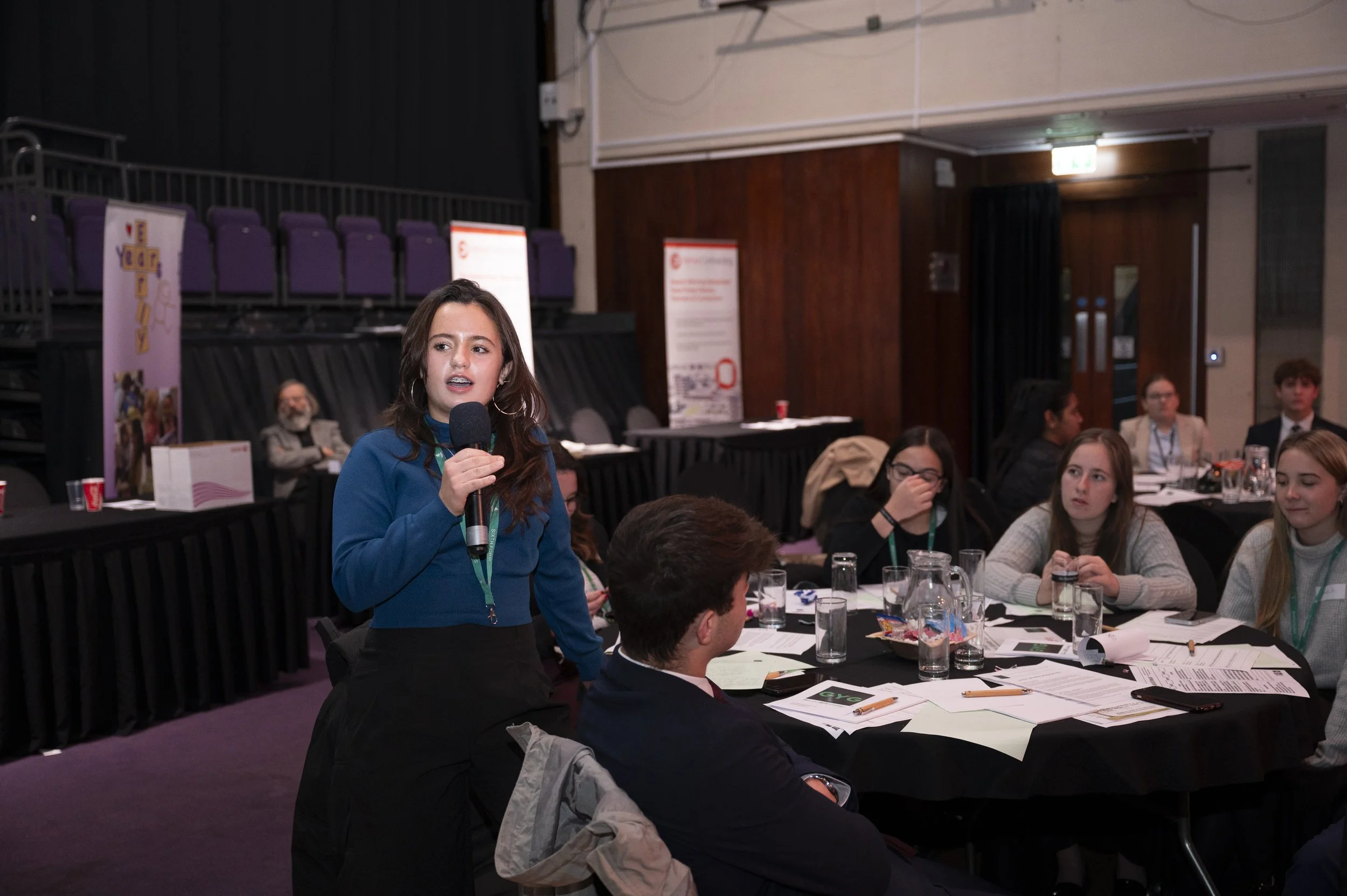 A woman speaking into a microphone at a conference or workshop. She stands near a table with several attendees sitting around it. Papers and glasses are scattered on the table. The background shows more attendees and conference banners.