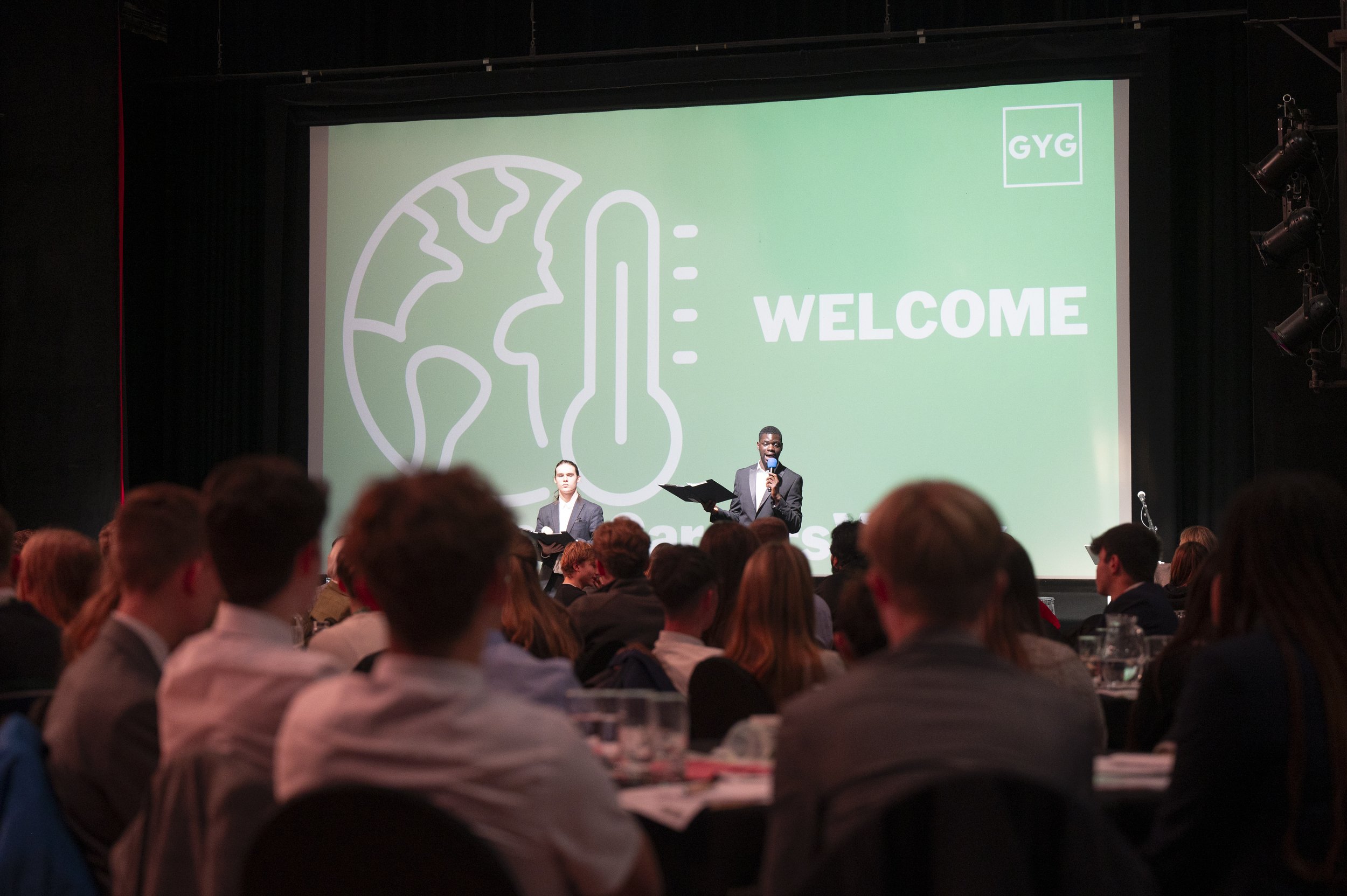 Conference scene with two speakers on stage, large welcome screen with earth and thermometer graphic, audience watching.