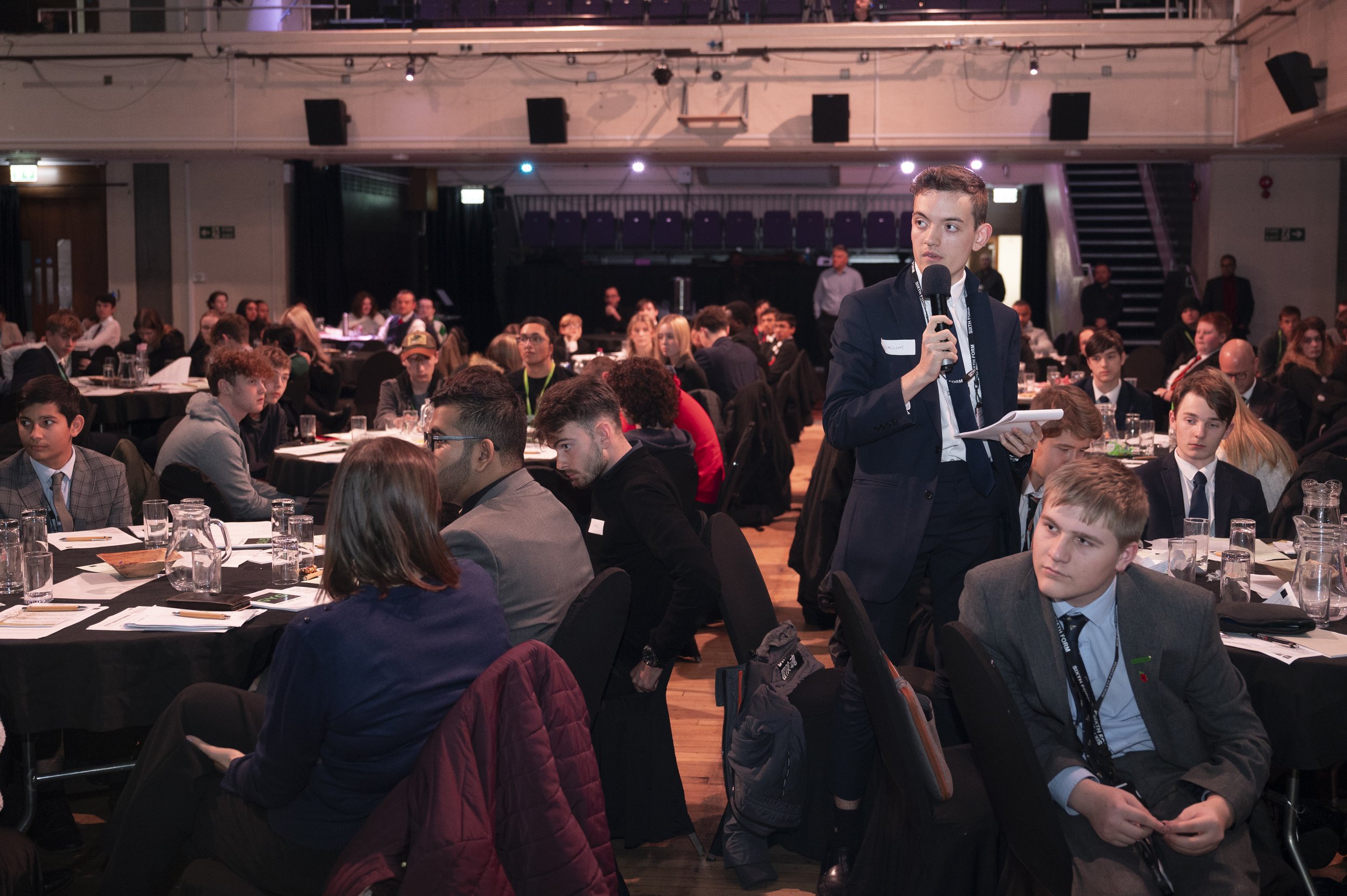 People sitting around tables in a conference setting with one person standing and speaking into a microphone.