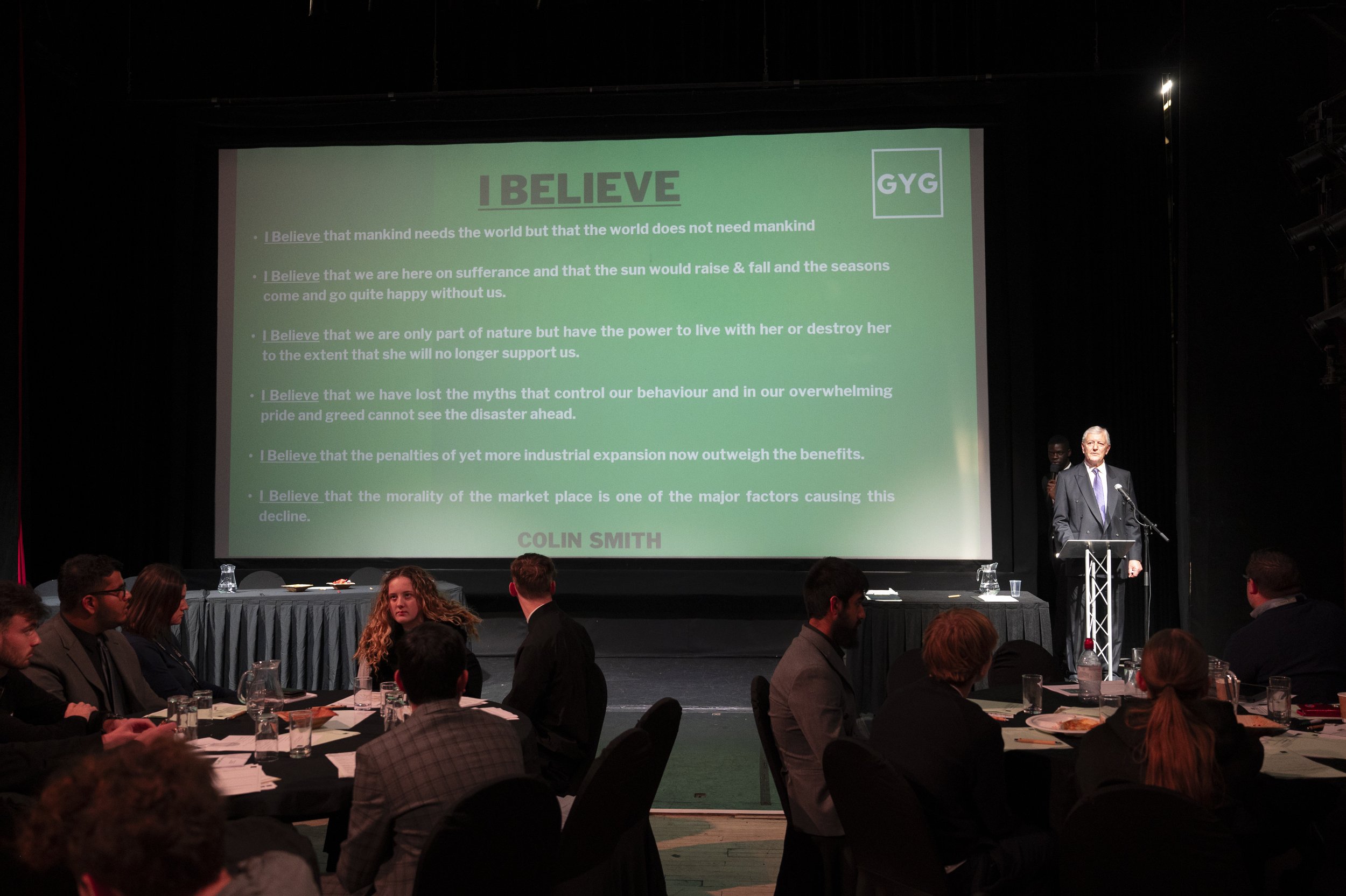 A speaker is giving a presentation on stage beside a large screen displaying text titled "I Believe," with a logo labeled "GYG." The text discusses philosophical views about mankind's relationship with nature and industrial expansion. In the foregrou