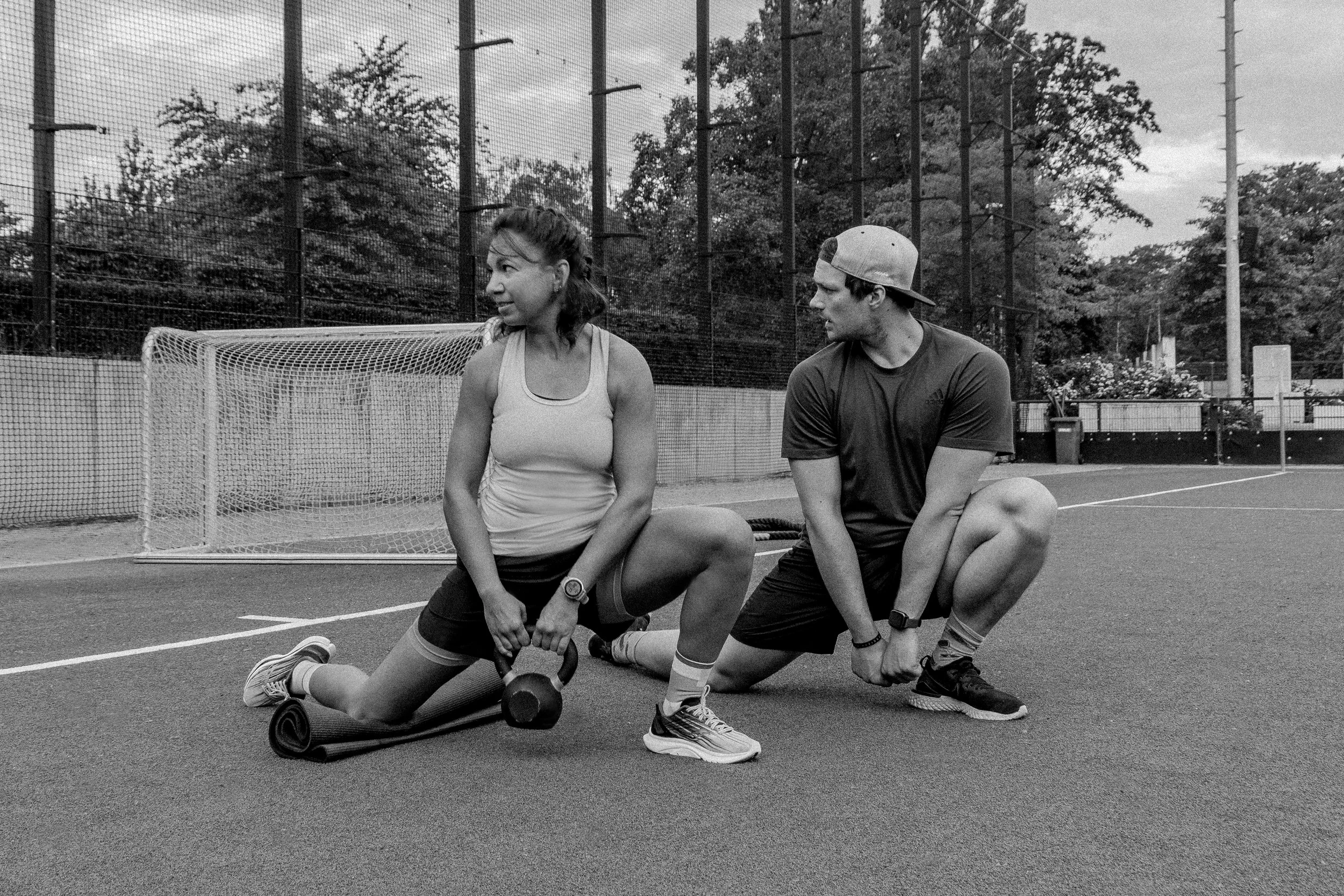 Zwei Menschen auf einem Sportplatz. Sie führen synchron eine Übung im Ausfallschritt aus. Dabei knien sie jeweils auf einer Matte.