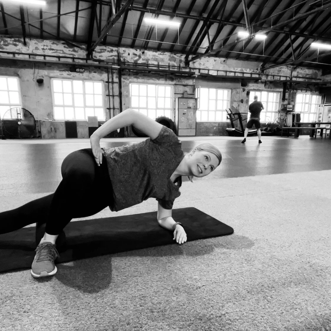 Frau macht eine Plank-Übung auf einer Matte in einem Fitnessstudio, im Hintergrund andere Personen beim Training, große Fenster und industrielle Gestaltung des Raumes.