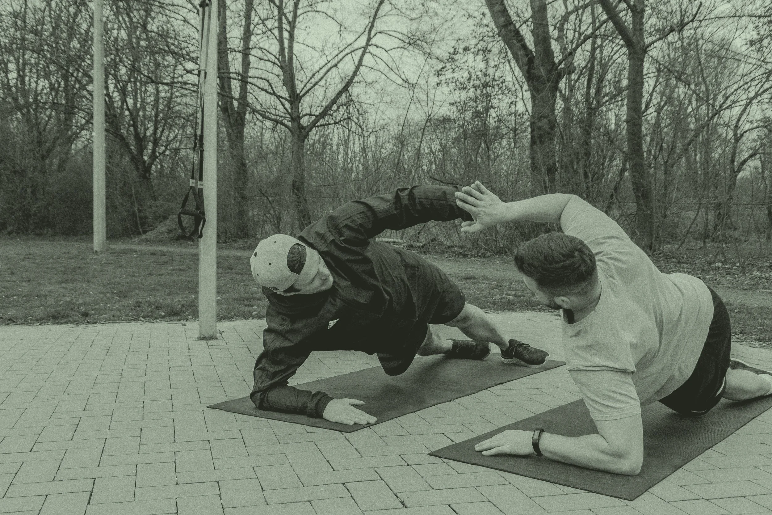 Zwei Männer trainieren in einem Park auf Yogamatten, einer führt eine Partnerübung aus, während der andere assistiert, im Hintergrund Bäume ohne Blätter.