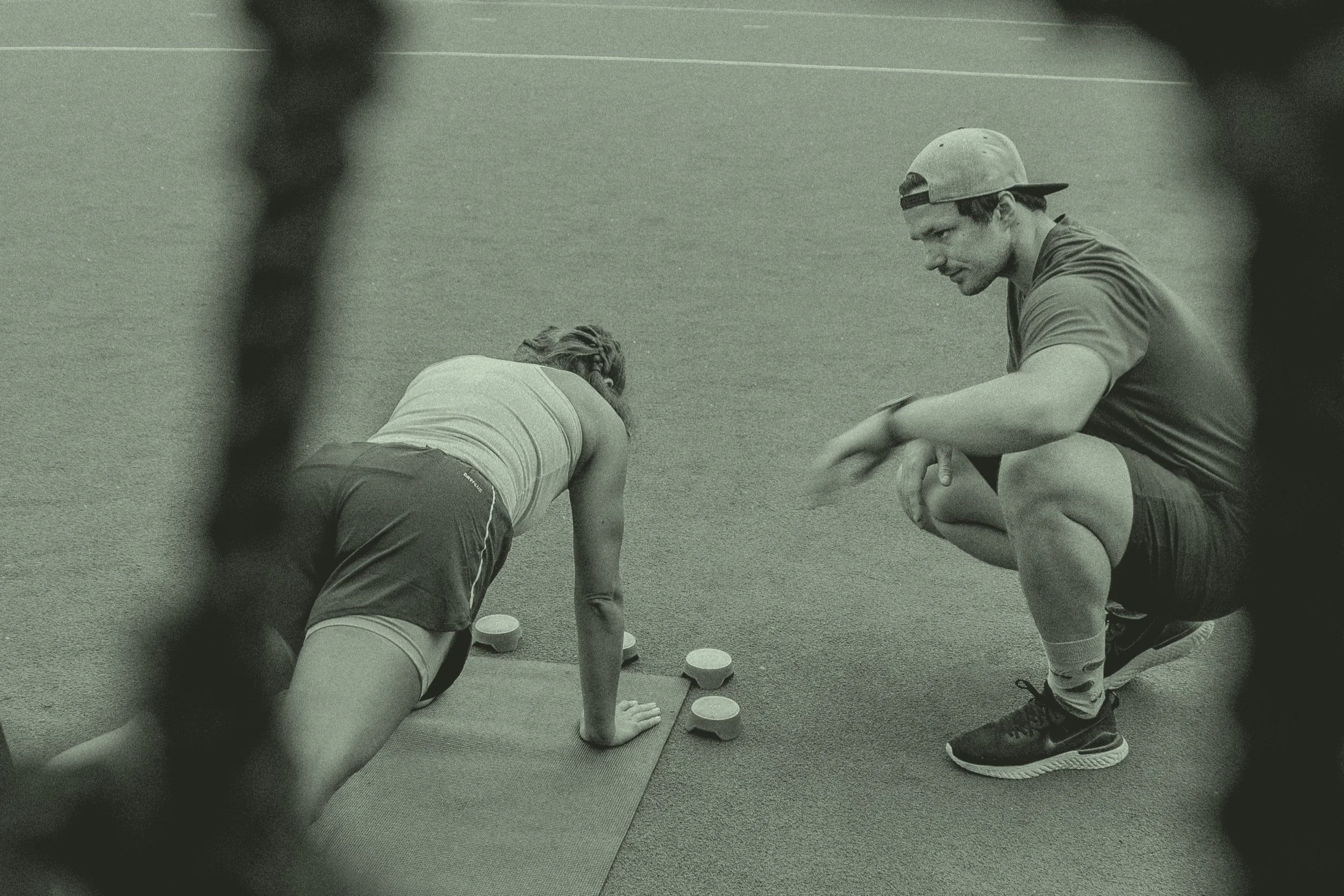 Ein Mann trägt eine Baseballkappe und sitzt auf dem Boden, während eine Frau in Sportkleidung in der Nähe auf allen Vieren auf dem Boden liegt. Die beiden befinden sich auf einer Turnmatte, und es sind kleine weiße Scheiben und Getränke neben ihnen.