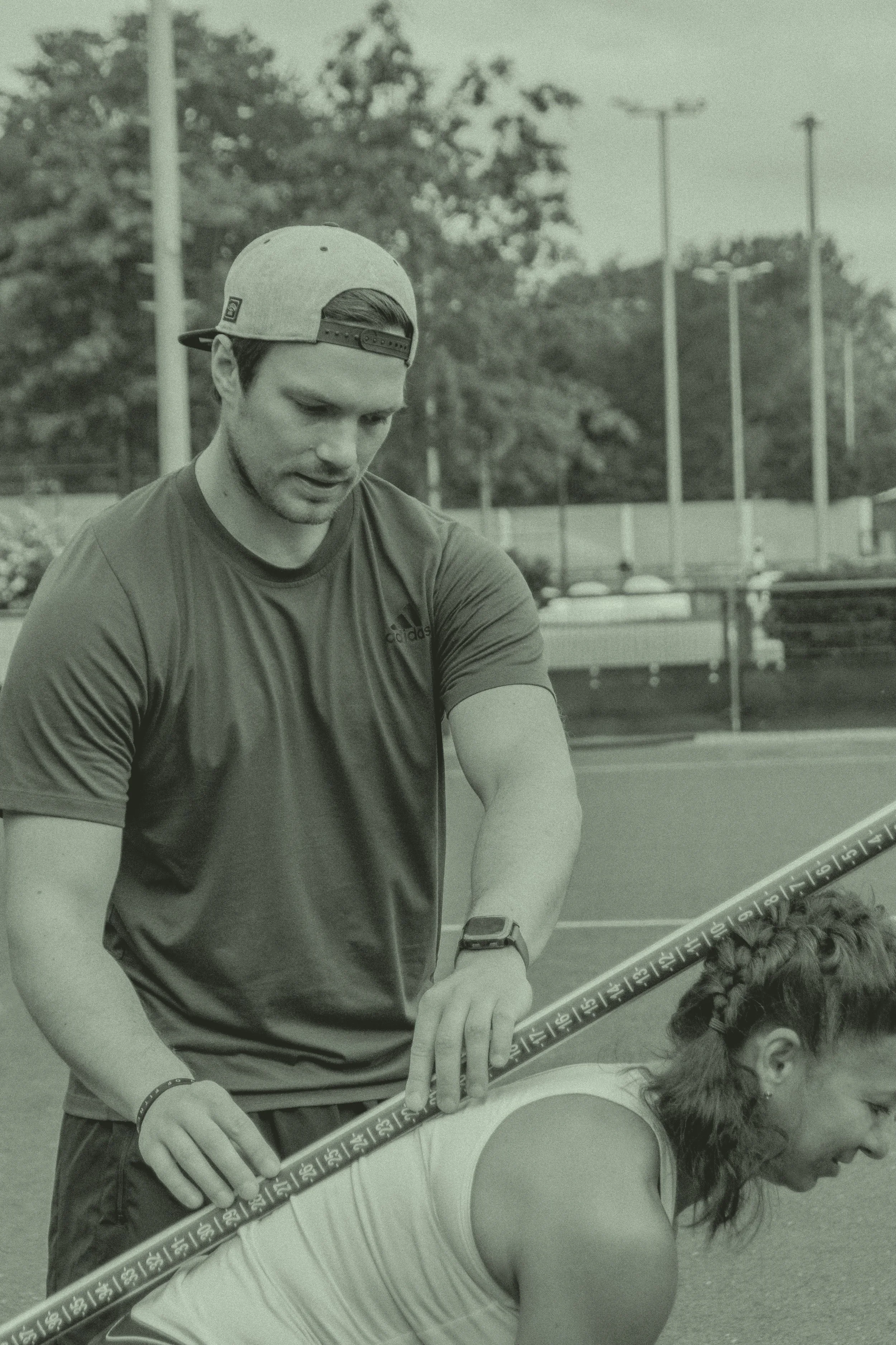 Ein junger Mann trägt eine Baseballkappe und ein Sport-T-Shirt, er steht im Freien bei einer Messung mit einem Messstab an einem Arm einer jungen Frau mit dunklen, lockigen Haaren, in einem Sportzentrum oder auf einem Sportplatz.