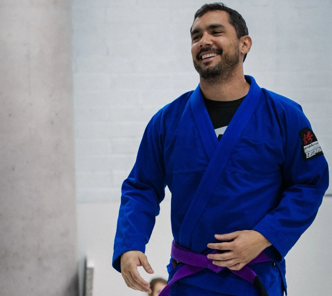 A man in a Brazilian jiu-jitsu gi smiling, wearing a purple belt, standing indoors.