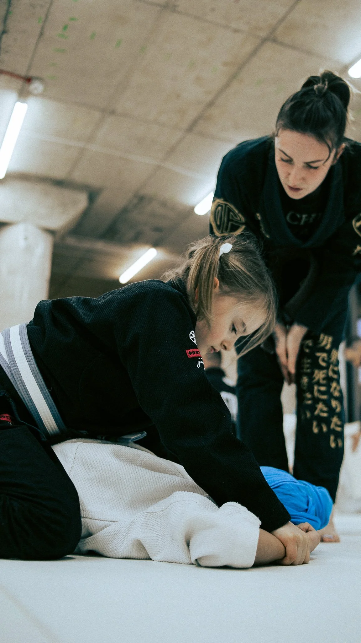 Young girl practicing Brazilian Jiu-Jitsu on a padded mat, with an instructor guiding her.