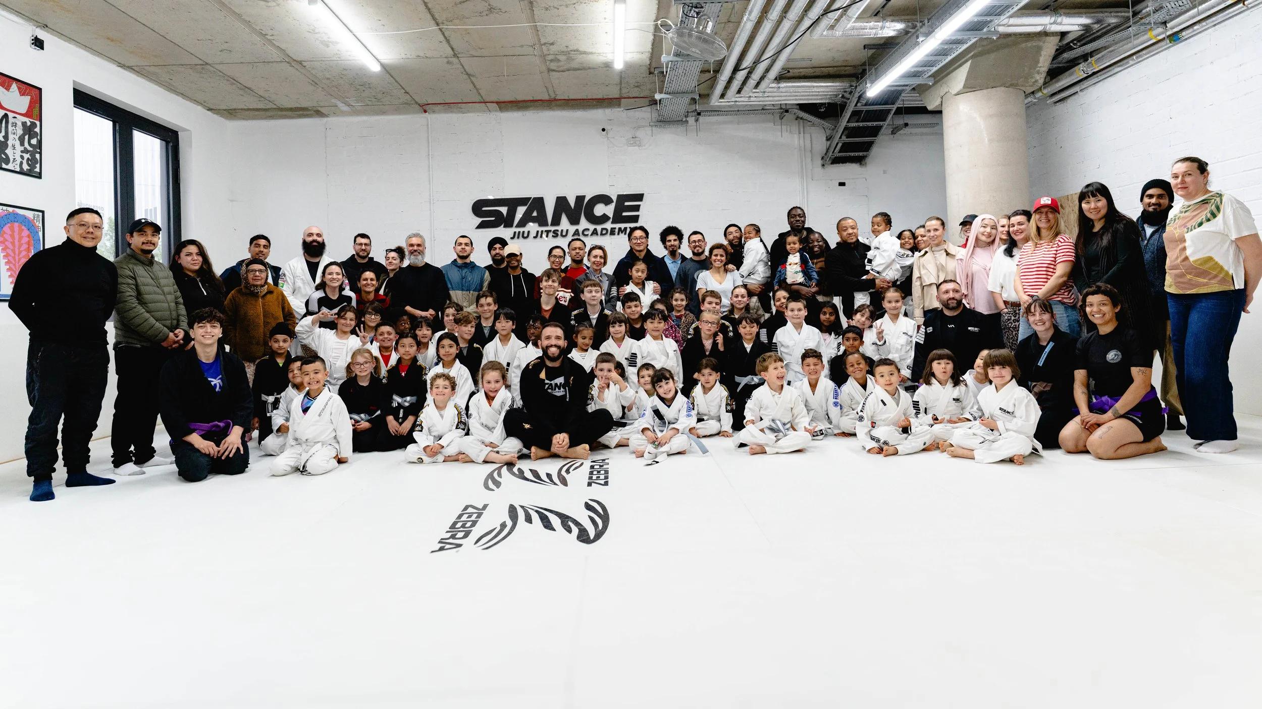A large group of people, including children in martial arts uniforms and adults, gathered in a martial arts training facility with the 'STANCE JIU JITSU ACADEMY' logo on the wall. The group is smiling and posing for a group photo on a white mat.