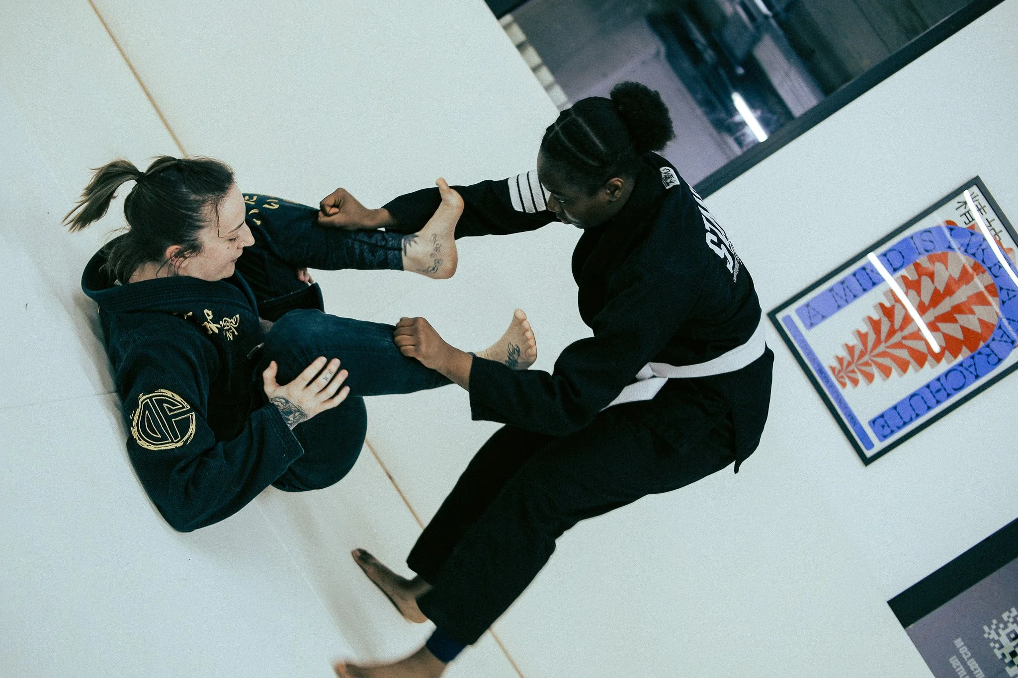 Two women practicing Brazilian Jiu-Jitsu on a mat, one sitting and the other kneeling, both in gis, in a gym with a framed martial arts poster on the wall.