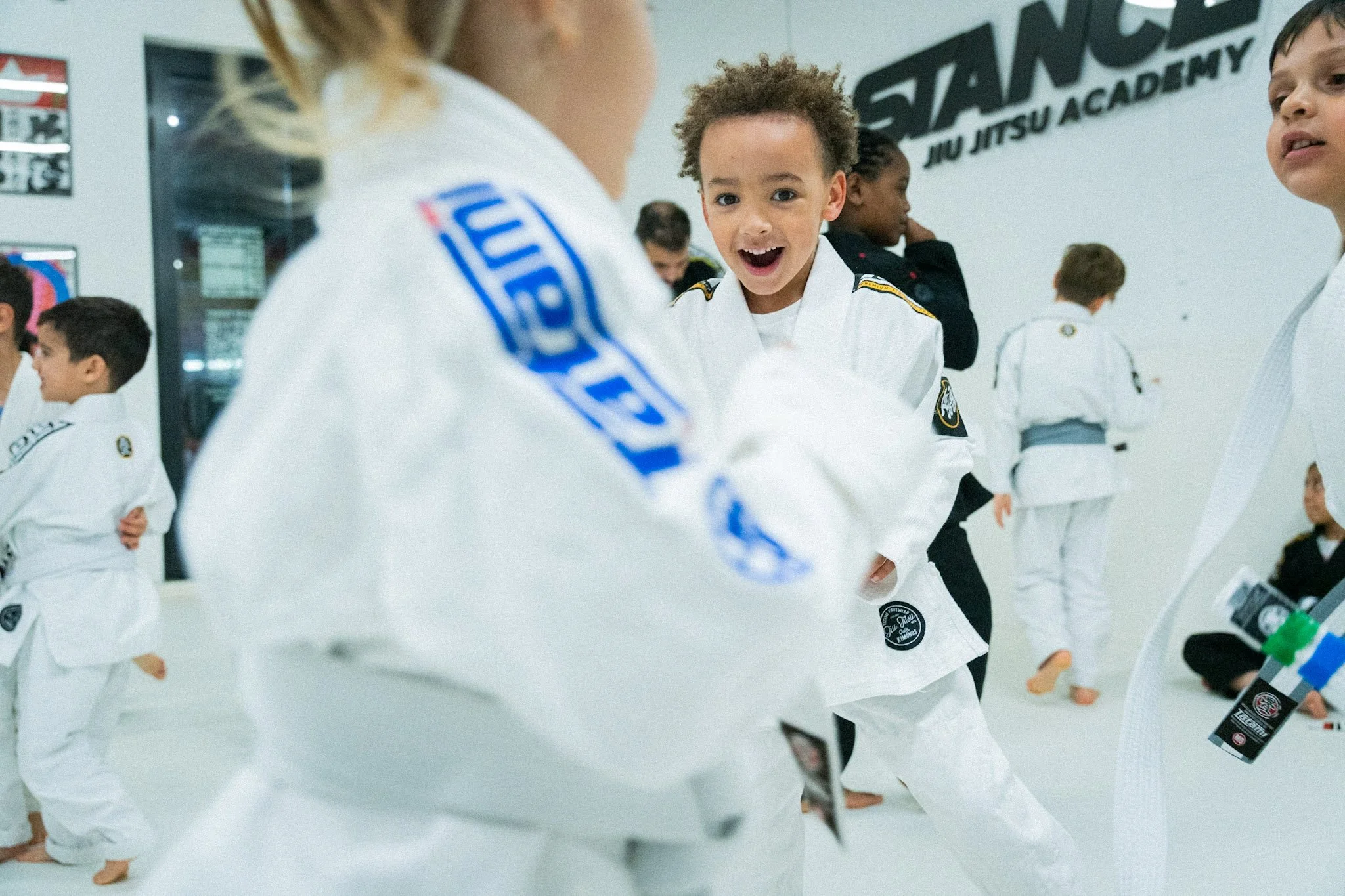 Young children practicing Brazilian Jiu-Jitsu in a martial arts gym, wearing white gis with black and colored patches. The kids are standing and engaging in training, with a smiling boy at the center.