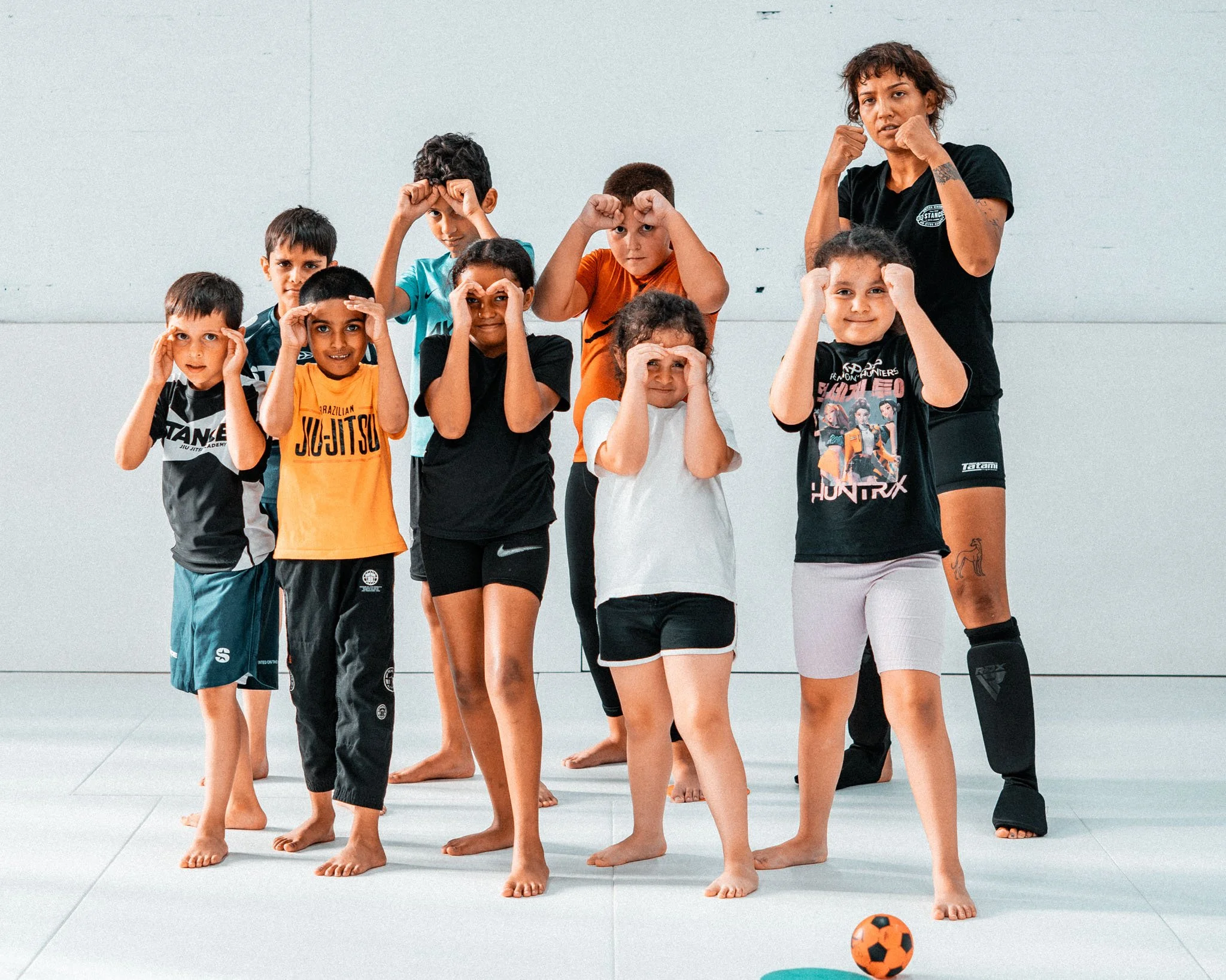 Five children in martial arts uniforms standing in a row inside a gym, with a sign that reads 'STANCE Jiu Jitsu Academy' in the background.