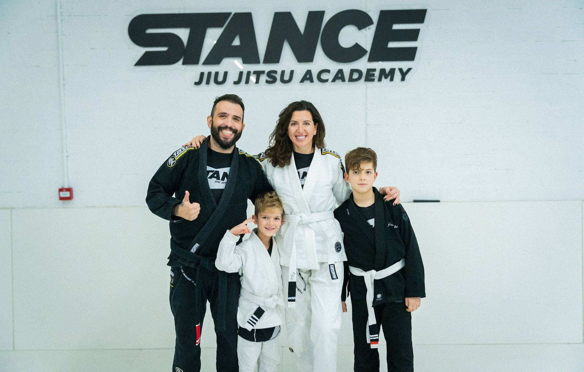 A smiling group photo of two Brazilian Jiu Jitsu instructors and three children wearing gi uniforms, standing in front of a wall with a large 'STANCE JIU JITSU ACADEMY' sign.