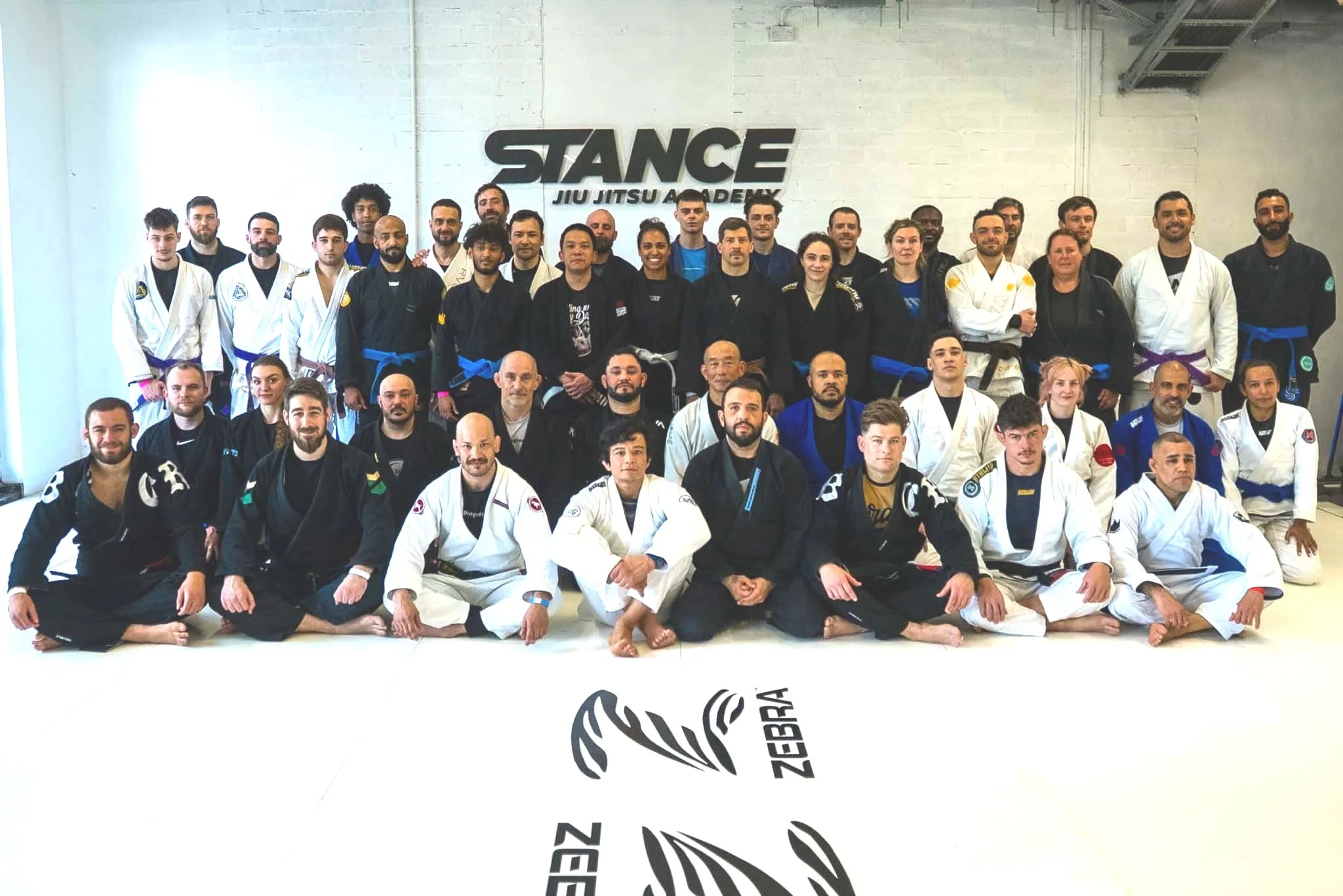 A large group of people in Brazilian Jiu Jitsu uniforms posing for a photo in a gym with a sign that says "STANCE JIU JITSU ACADEMY" on the wall.