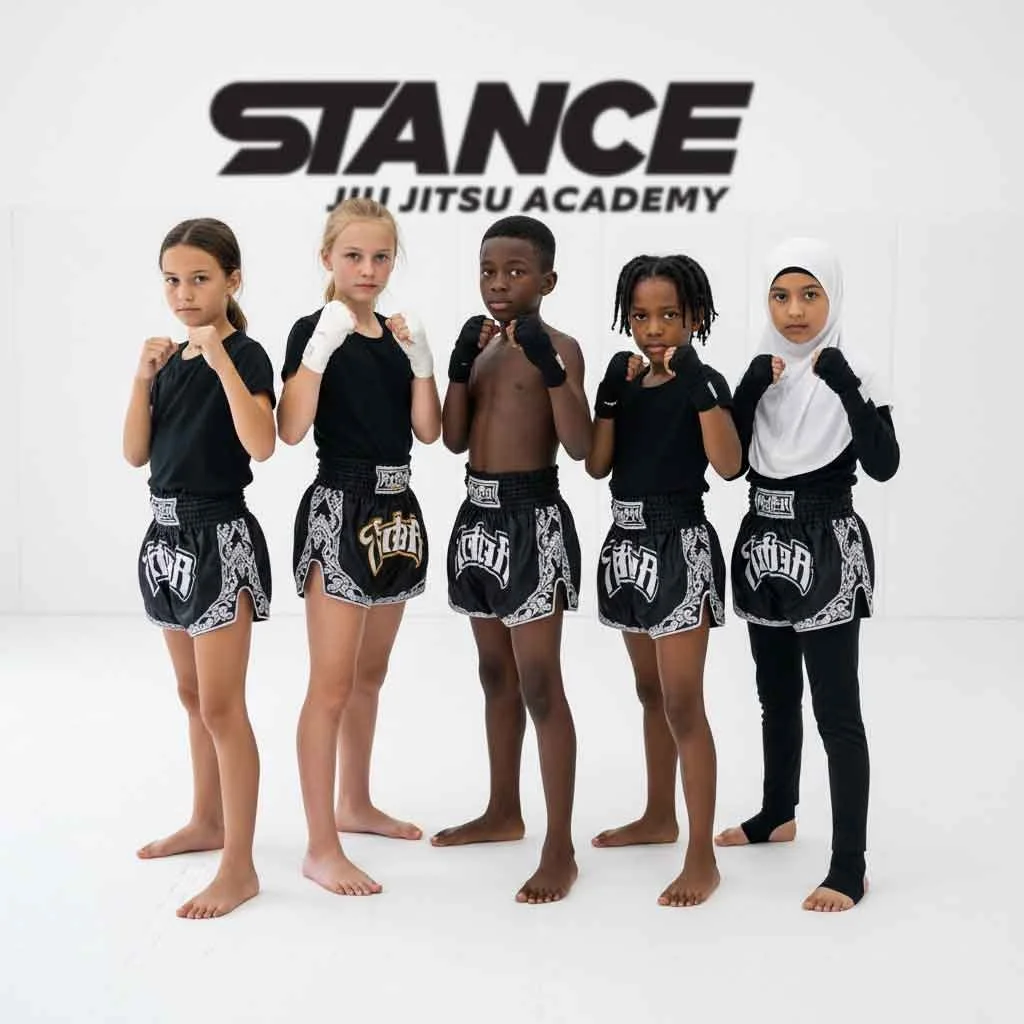 Five children in martial arts uniforms standing in a row inside a gym, with a sign that reads 'STANCE Jiu Jitsu Academy' in the background.