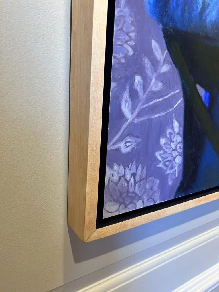 Close-up of a corner of a framed painting with a light wooden frame, green and purple abstract floral design, hung on a white wall near a baseboard.