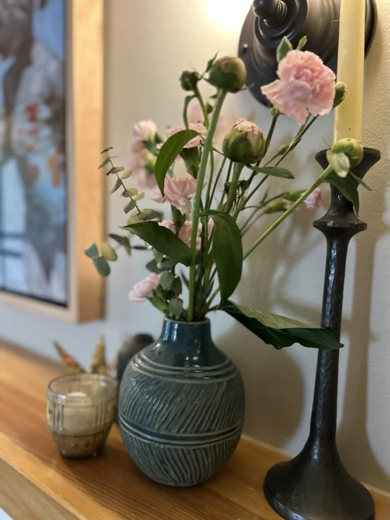 A blue vase with pink flowers and greenery, a small candle holder, and a tall black candle holder on a wooden surface against a wall.