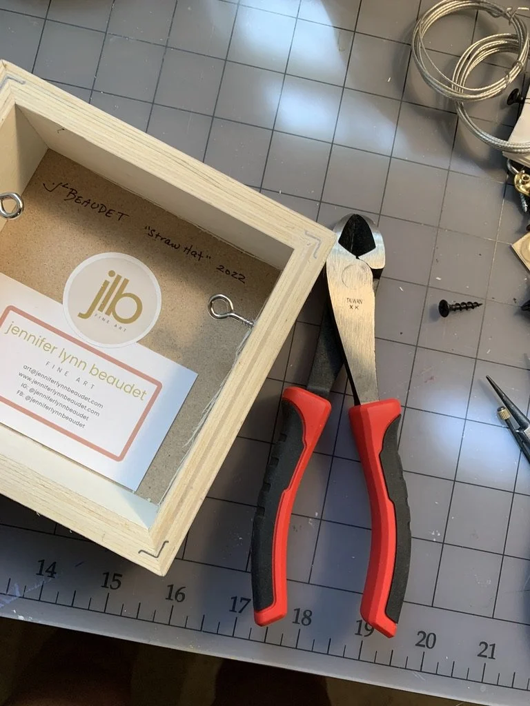 A worktable with a small wooden box labeled 'V. Beaudet 'Straw Hat' 2022', a folded business card for Jennifer Lynn Beaudet Fine Art, and a pair of pliers with red handles, surrounded by various tools and cords.