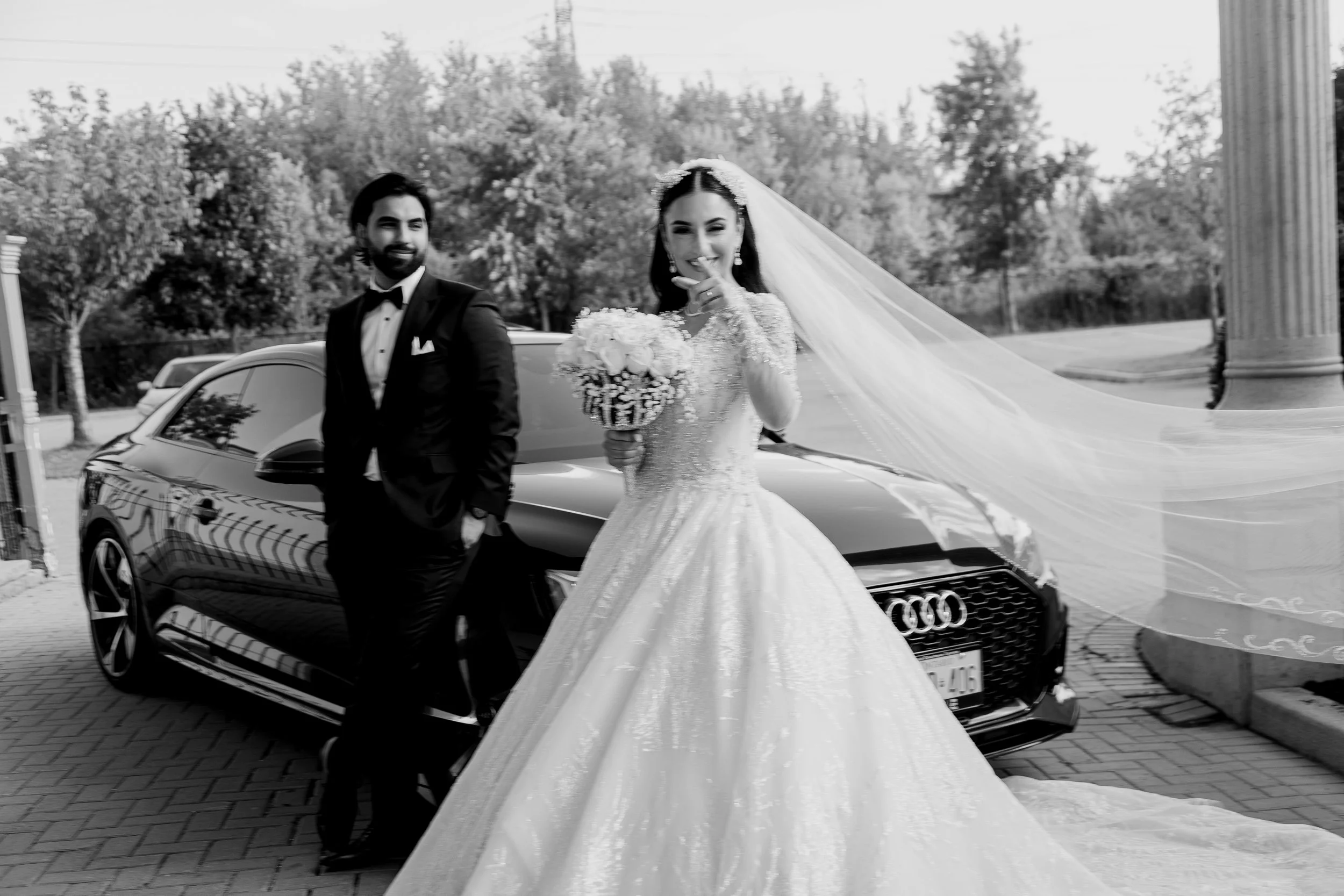 Black and white photo of a bride and groom standing beside a black luxury car with a scenic background of trees and a column, likely at a wedding.
