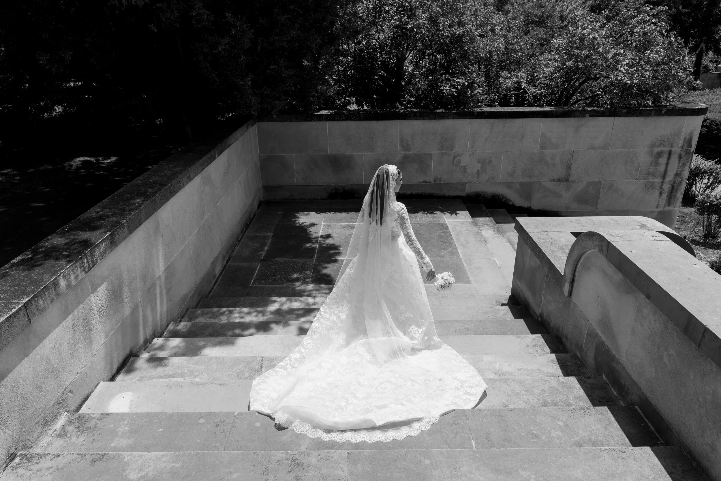 A bride walking down stairs holding a bouquet, wearing a long lace wedding dress and veil outdoors, with trees and a stone wall in the background.