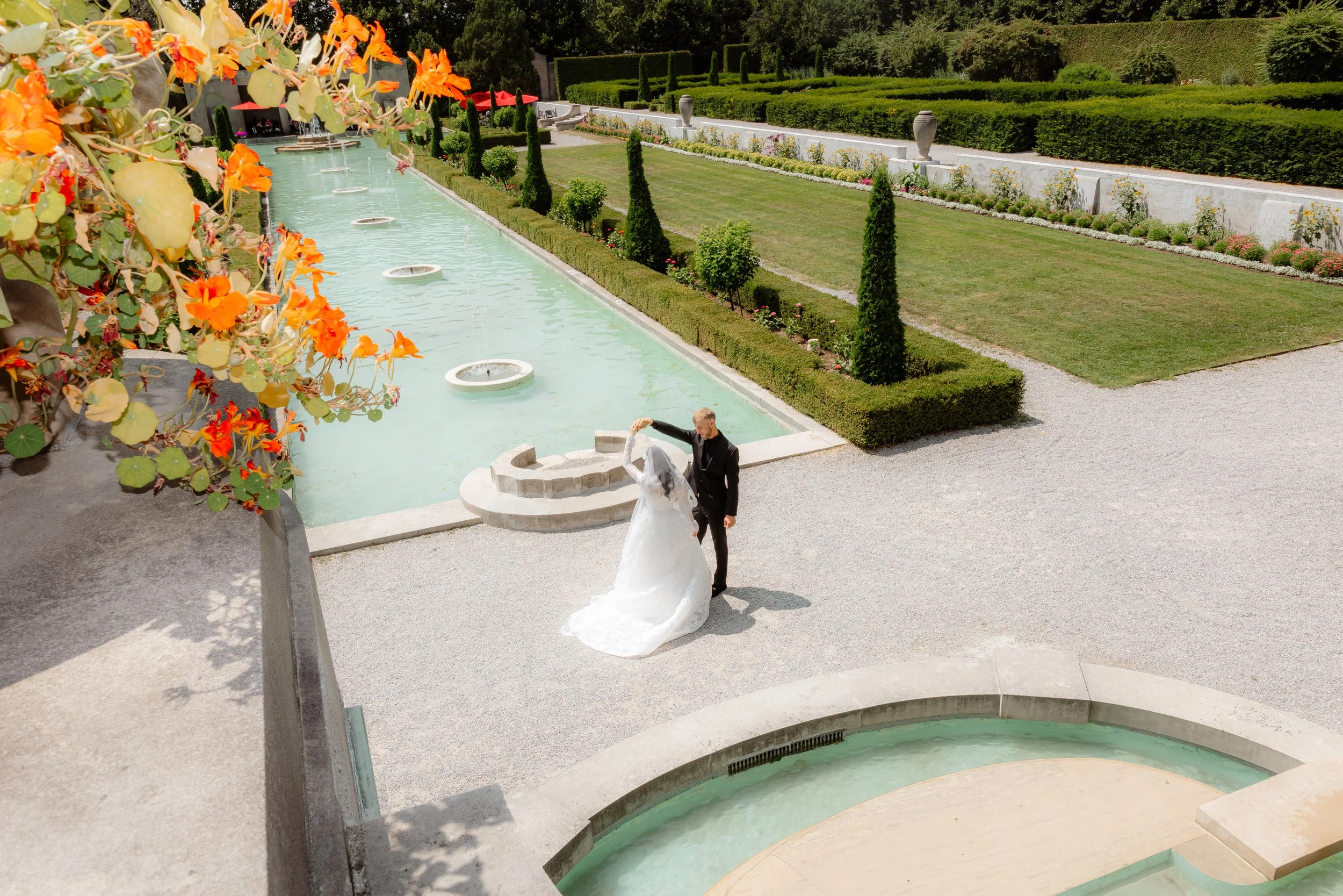 A bride and groom dancing outdoors near a rectangular fountain with water jets, in a landscaped garden with trimmed bushes, flowers, and trees.