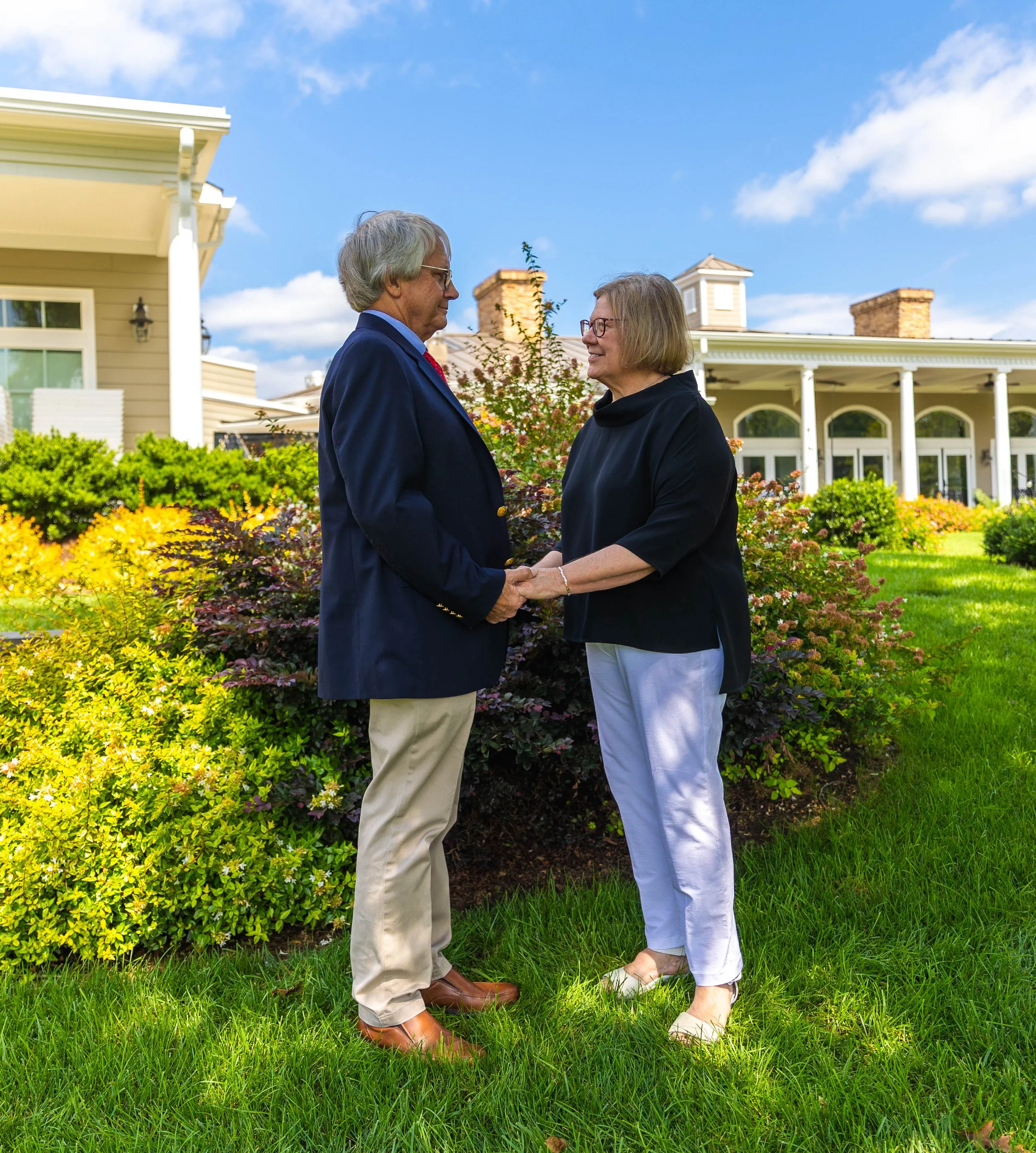 A man and a woman holding hands and looking at each other in a garden in front of a house with white columns and a porch, under a blue sky with clouds.