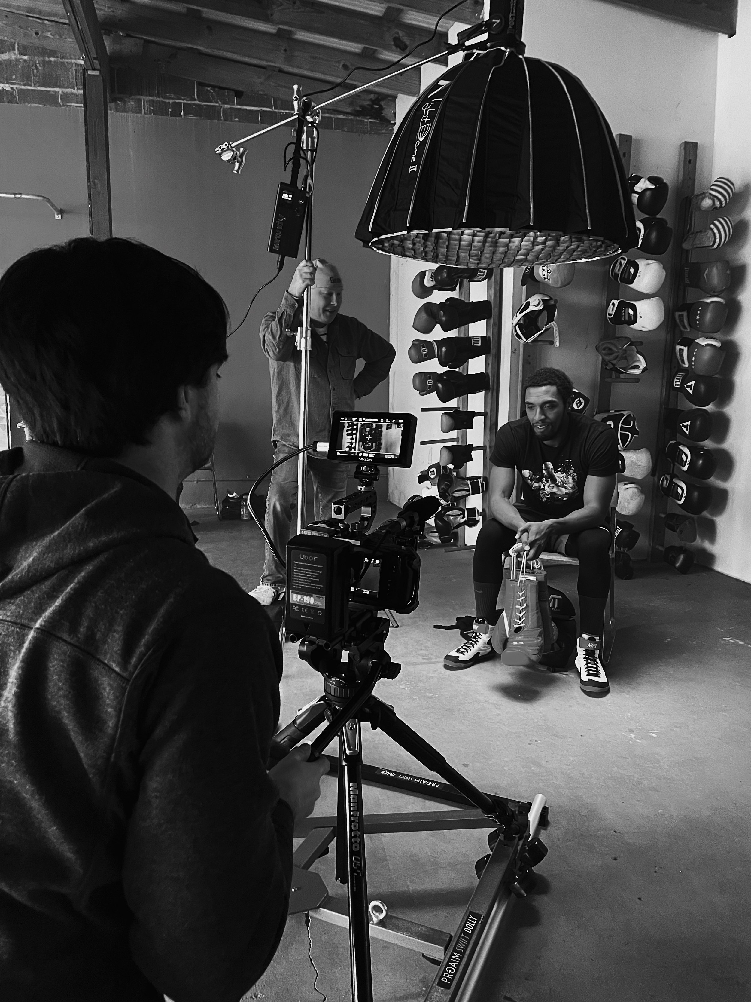 Filming of a boxing or training video in a gym with a man seated holding a pair of boxing gloves, a camera operator recording, lights, and boxing gloves on the wall.
