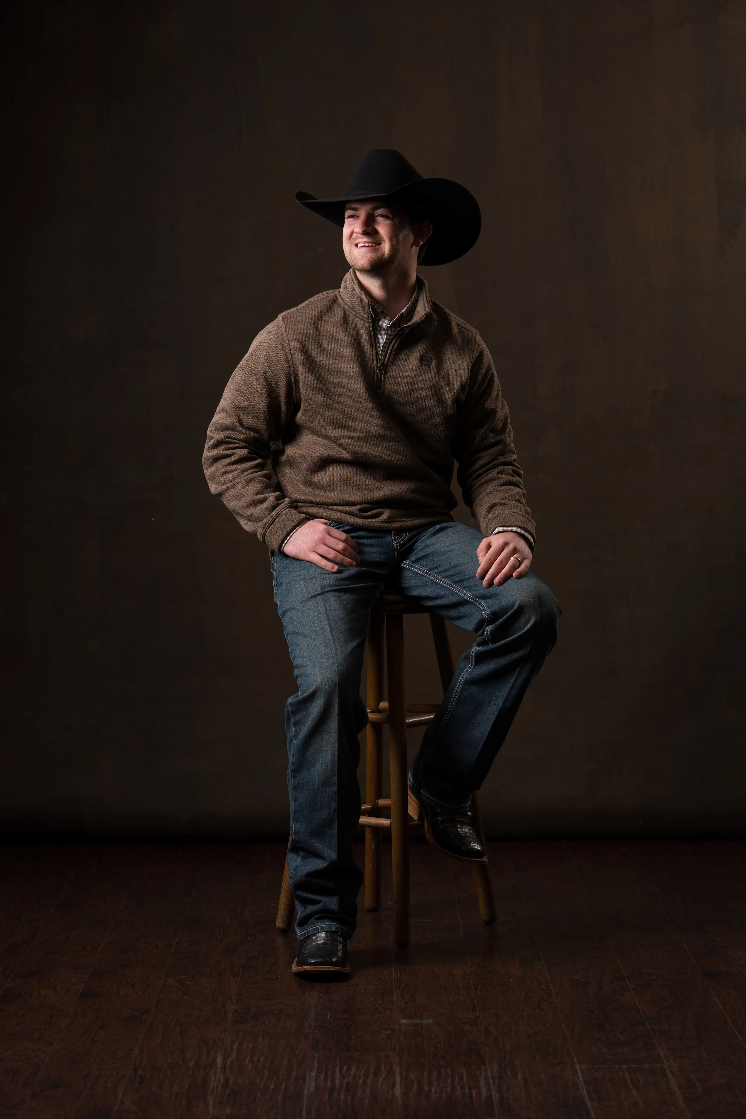 A man wearing a cowboy hat, a brown sweater, jeans, and cowboy boots sitting on a wooden stool, smiling, in a studio setting.