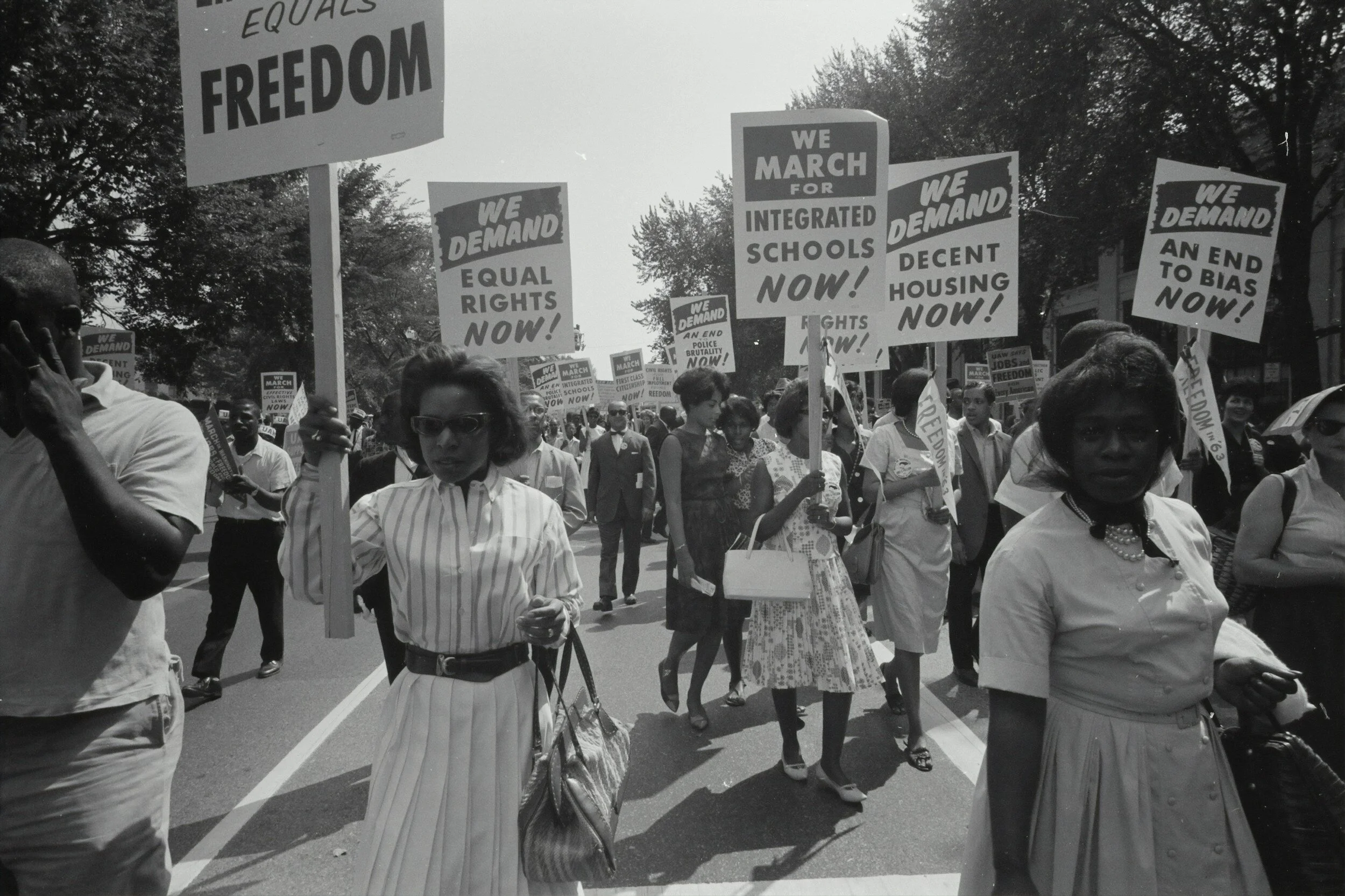 Black and white photograph of a civil rights march with people holding signs demanding equal rights, integrated schools, decent housing, and an end to bias, featuring diverse participants.