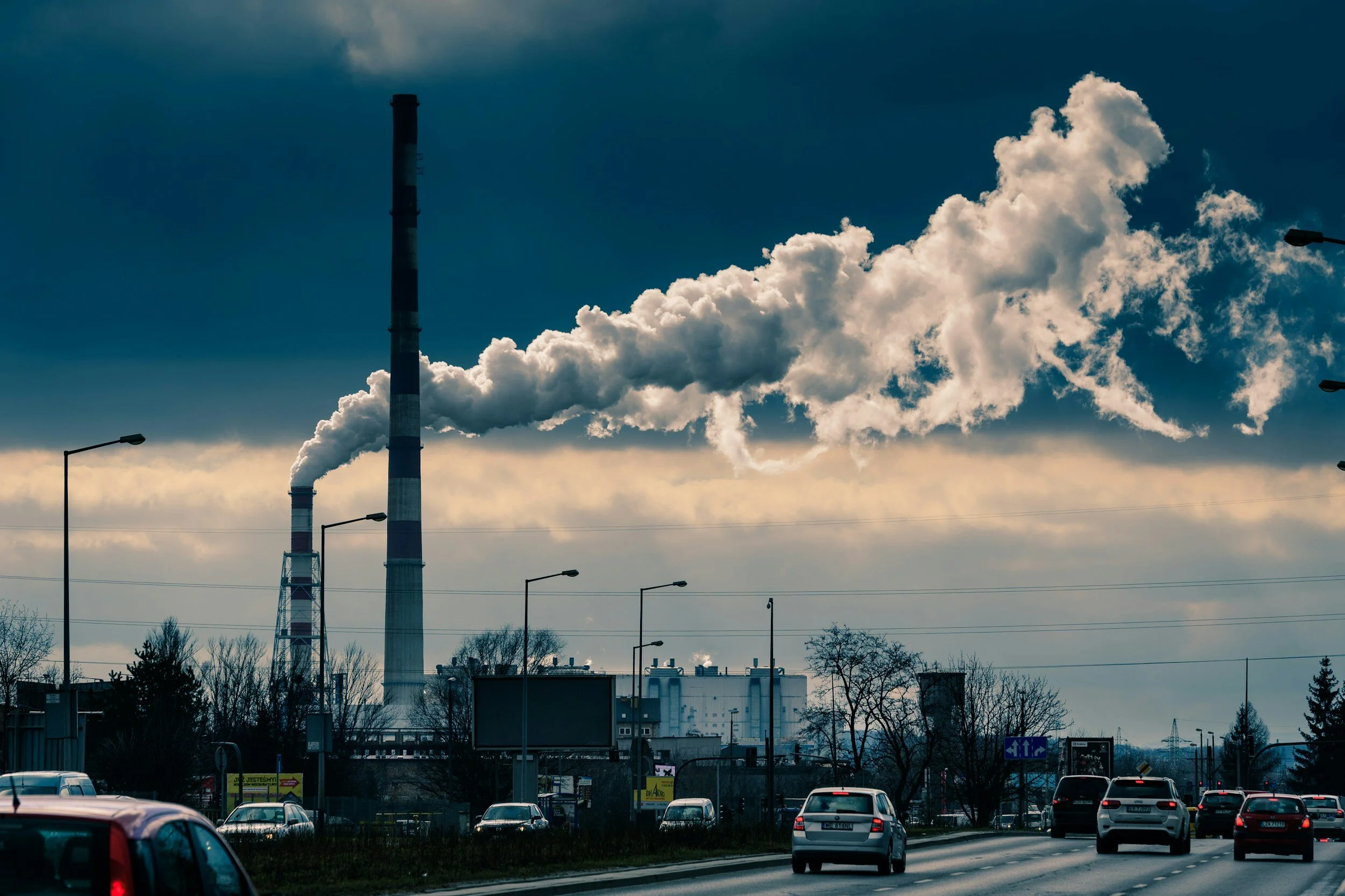 Cars driving on a road with a factory in the background emitting large plumes of smoke into a cloudy sky.