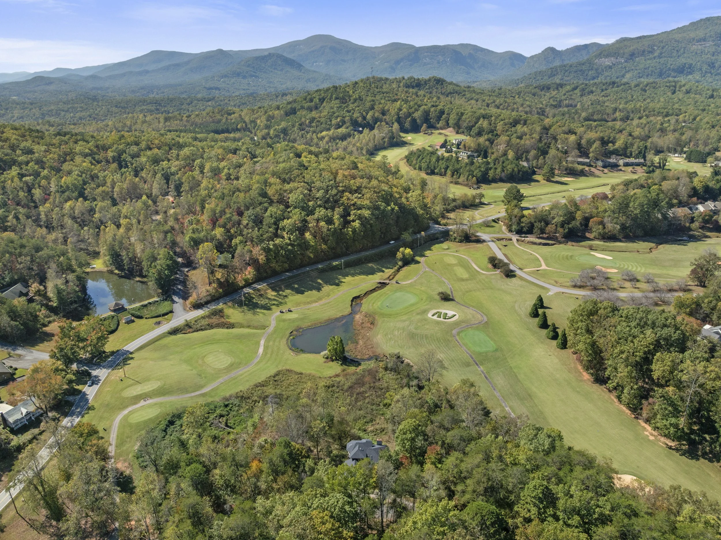 WNC drone photography. Aerial view of a golf course situated in a lush, heavily wooded landscape with rolling hills and mountains in the background.