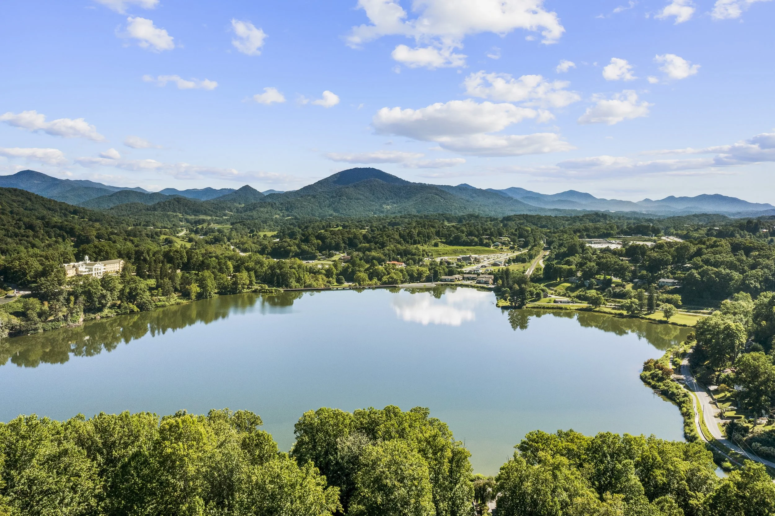 A large, calm lake surrounded by lush green trees and hills, with a mountain range in the background and partly cloudy sky.